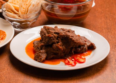 Close-up of savoury beef rendang with chili and sides on a wooden table.