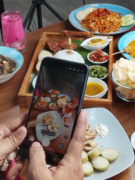Close-up of a traditional Indonesian meal being photographed on a smartphone, featuring satay and colorful dishes.