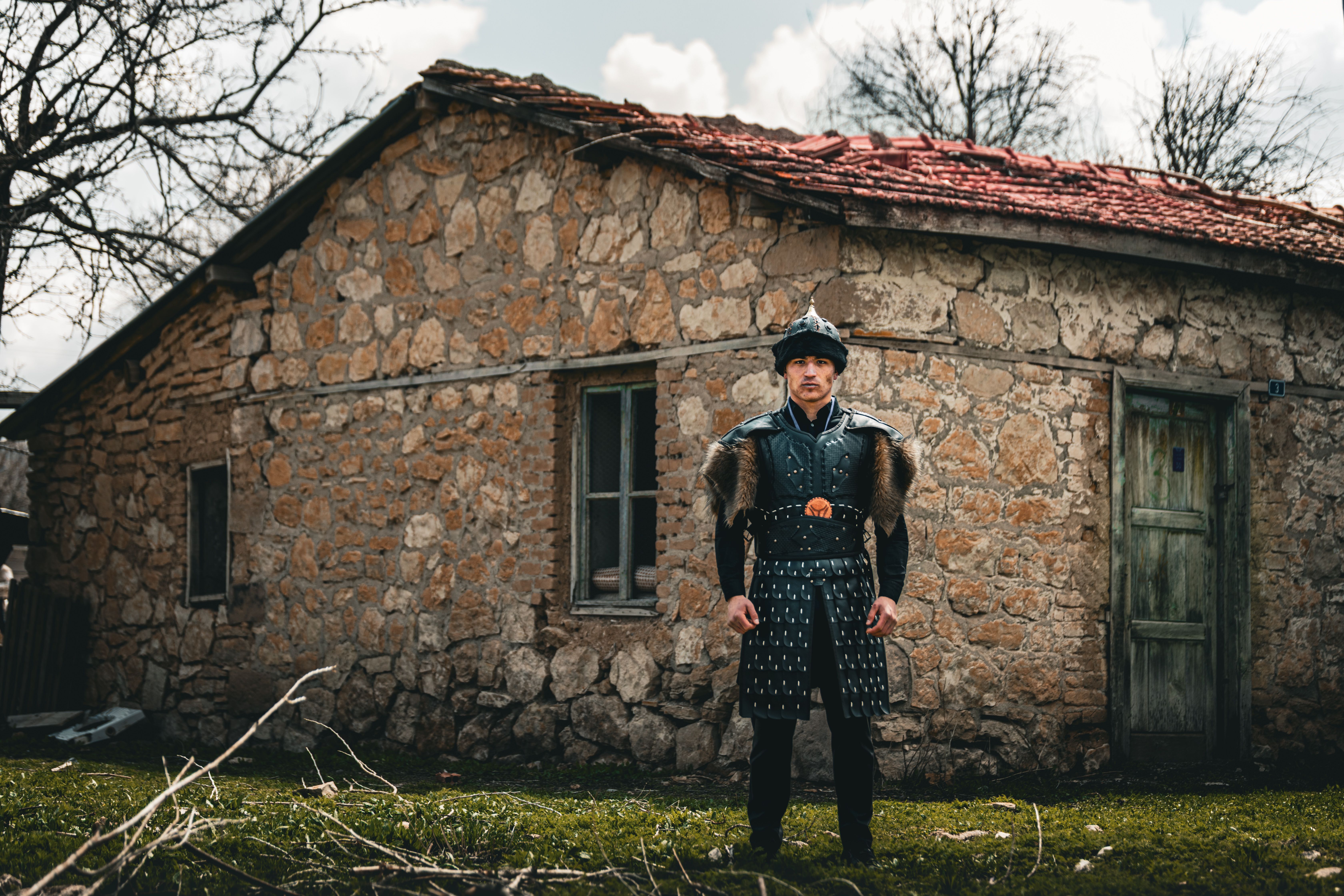 A medieval warrior in armor stands in front of a rustic stone cottage, evoking a historical atmosphere.
