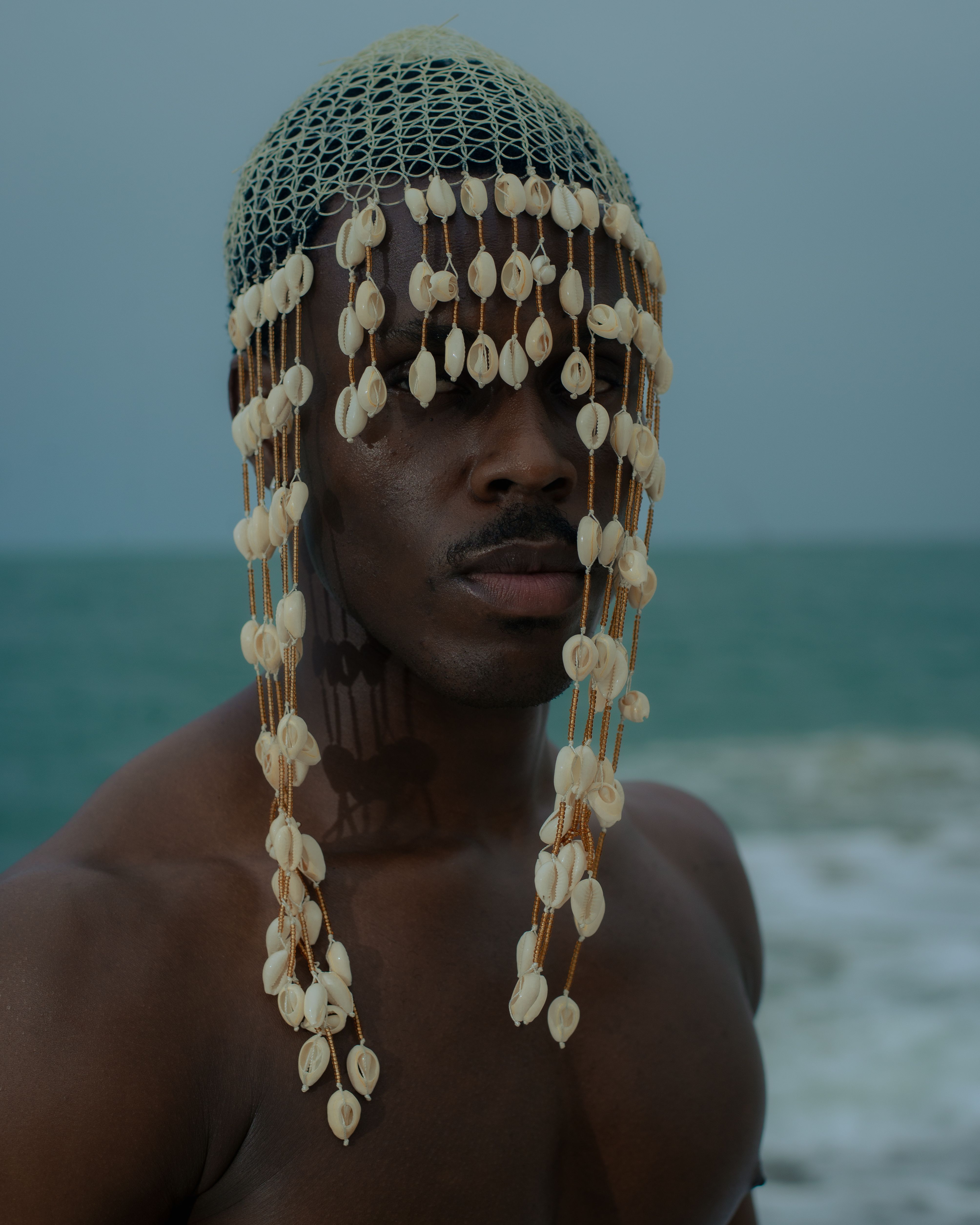 An artistic portrait of a man wearing a unique shell headpiece by the ocean in Cotonou, Benin.