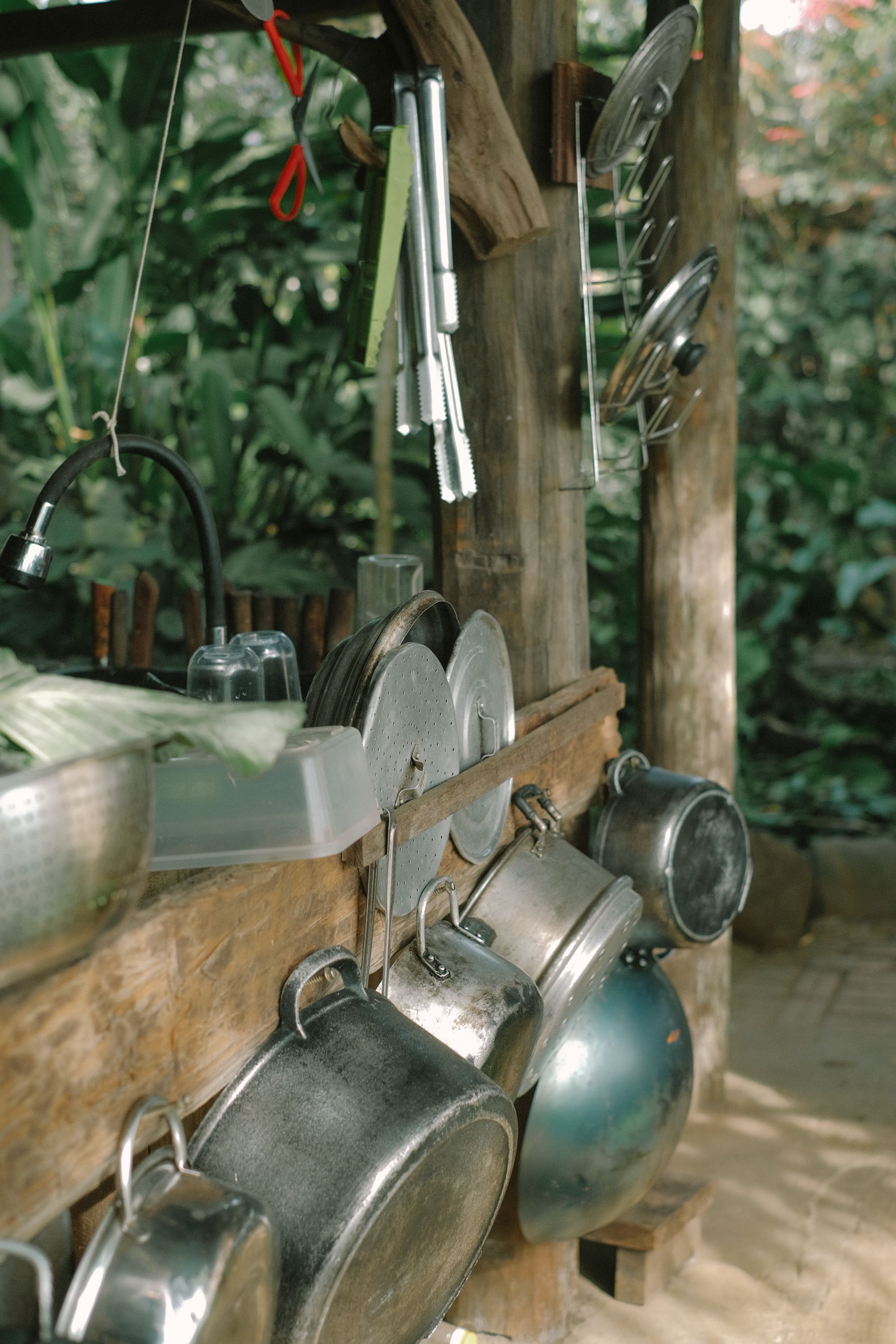 Rustic outdoor kitchen setup with hanging pots and pans against lush greenery.