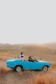 Two women enjoying a road trip in a retro blue convertible on a scenic hillside at sunset.
