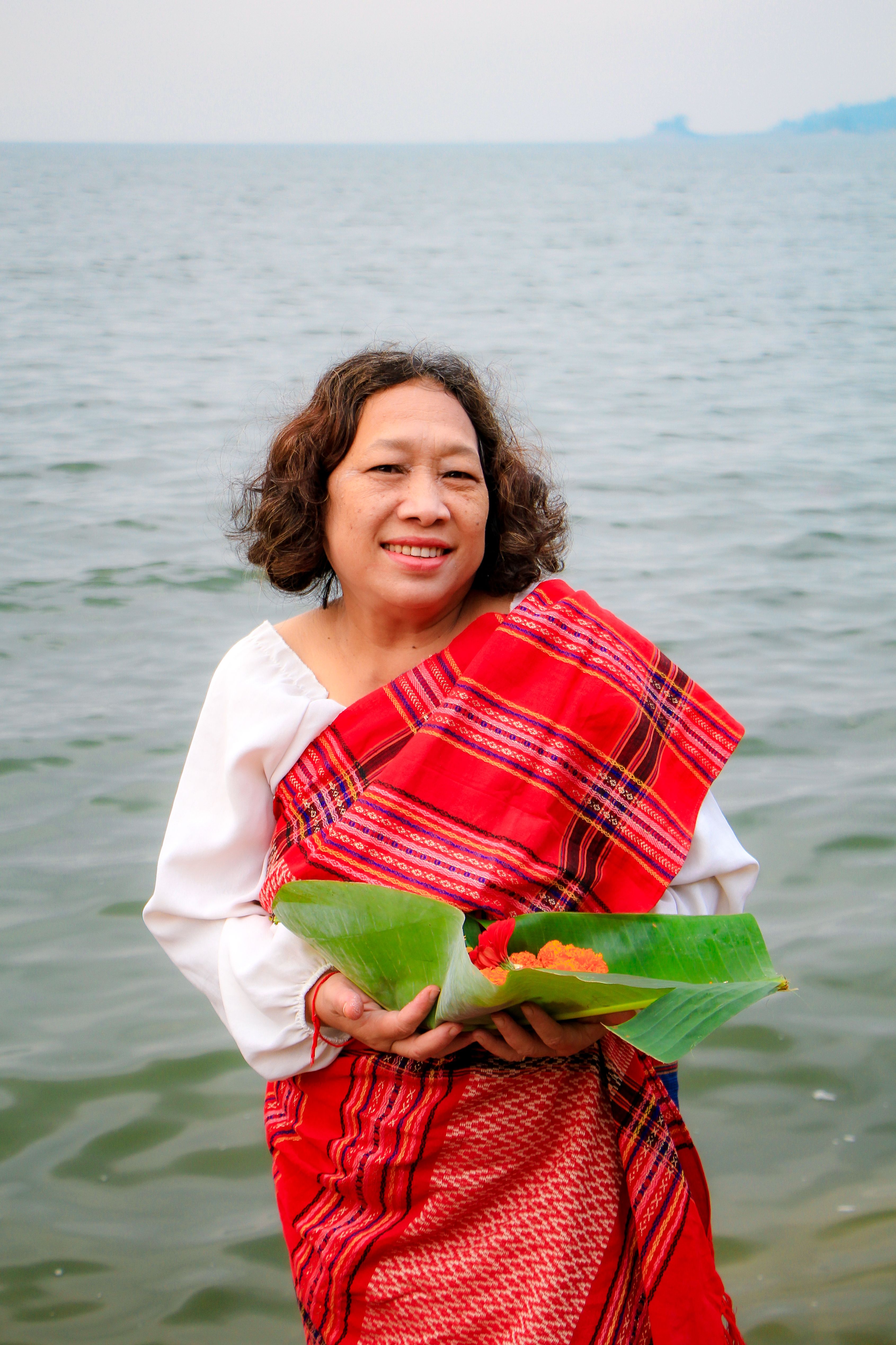 A woman in traditional attire holds a leaf with flowers by the Rangamati river.