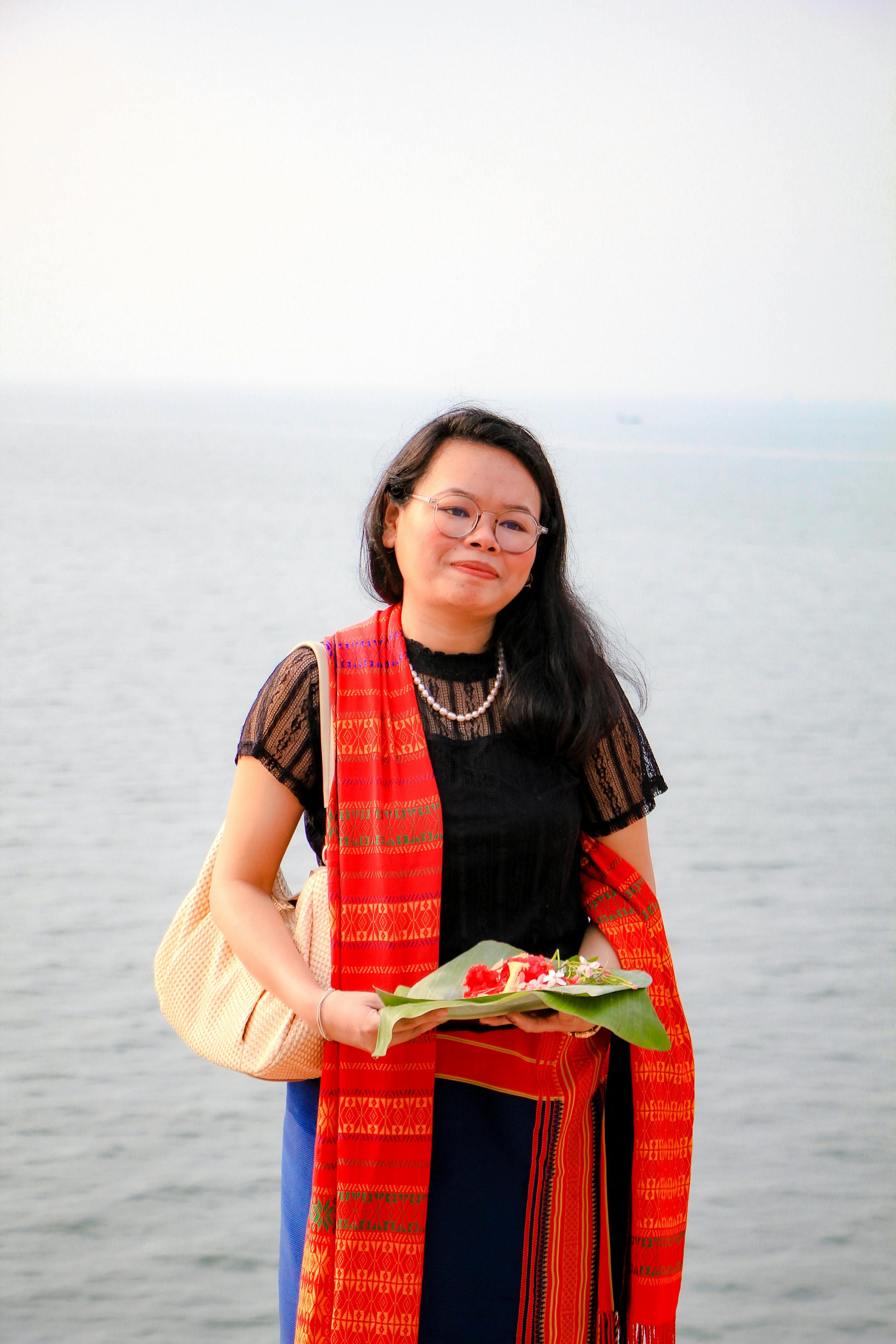 A woman wearing traditional Chakma clothing stands by the river holding a flower offering, celebrating cultural heritage.