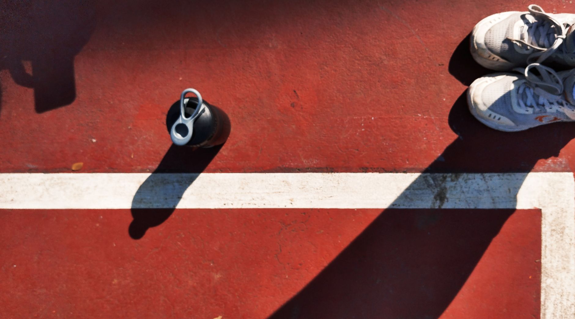 Overhead view of white sneakers and a water bottle on a red sports court with a dividing line.