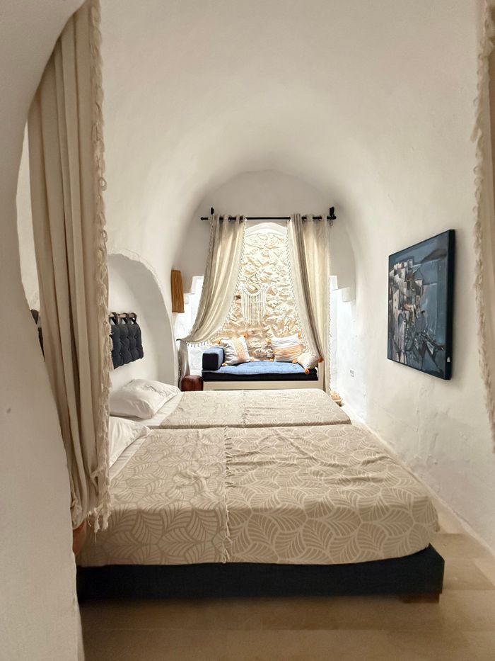 A long white vaulted room with a double bed and a curtained window seat at the far end