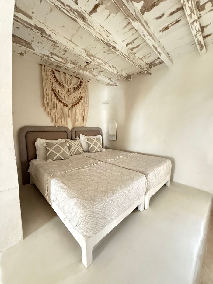 A whitewashed vaulted room with two single beds under crocheted covers and a woven wall hanging
