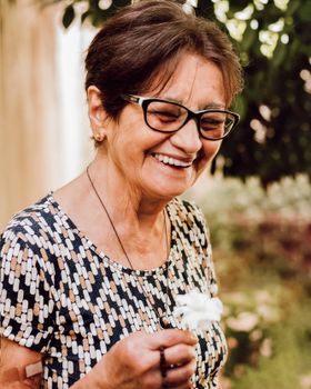 Senior woman with glasses, smiling joyfully outdoors while holding a flower, exuding happiness