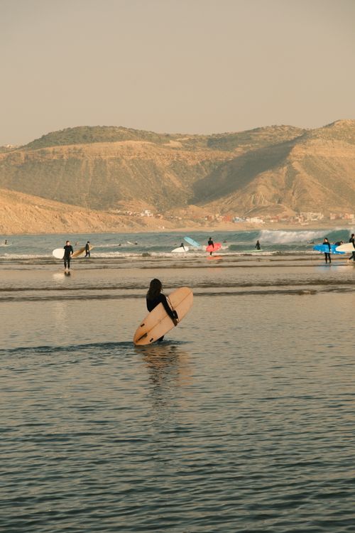 Free A surfer with a board approaches waves at a scenic beach with hills in the background. Stock Photo