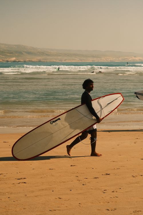 Free A surfer carrying a longboard walks along a tranquil sandy beach by the ocean. Stock Photo