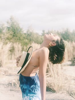 Young topless woman in shorts enjoying the sun, holding a hat in a sunny outdoor setting.