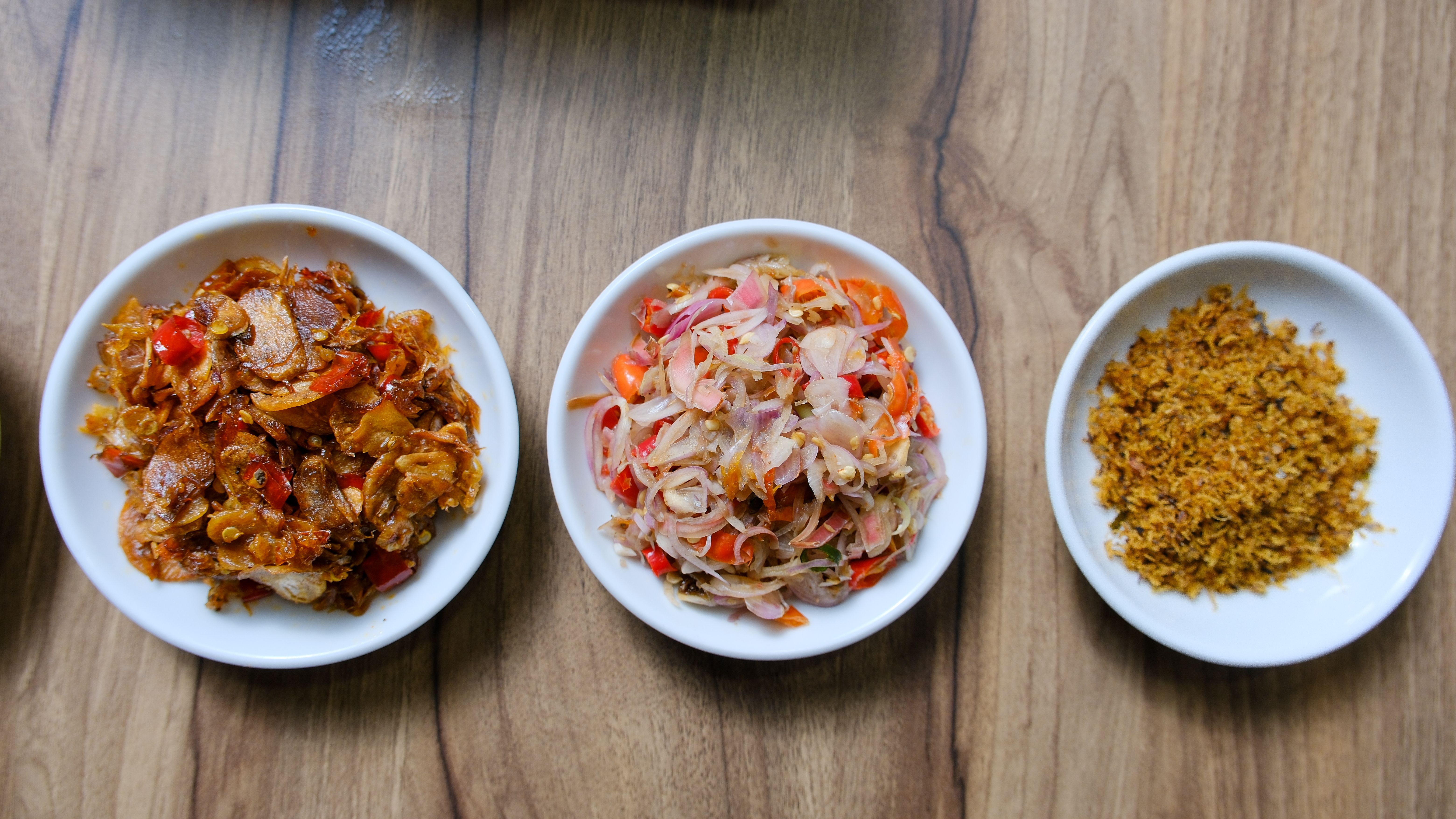 Top-down view of three traditional Indonesian side dishes served on white plates against a wooden table.