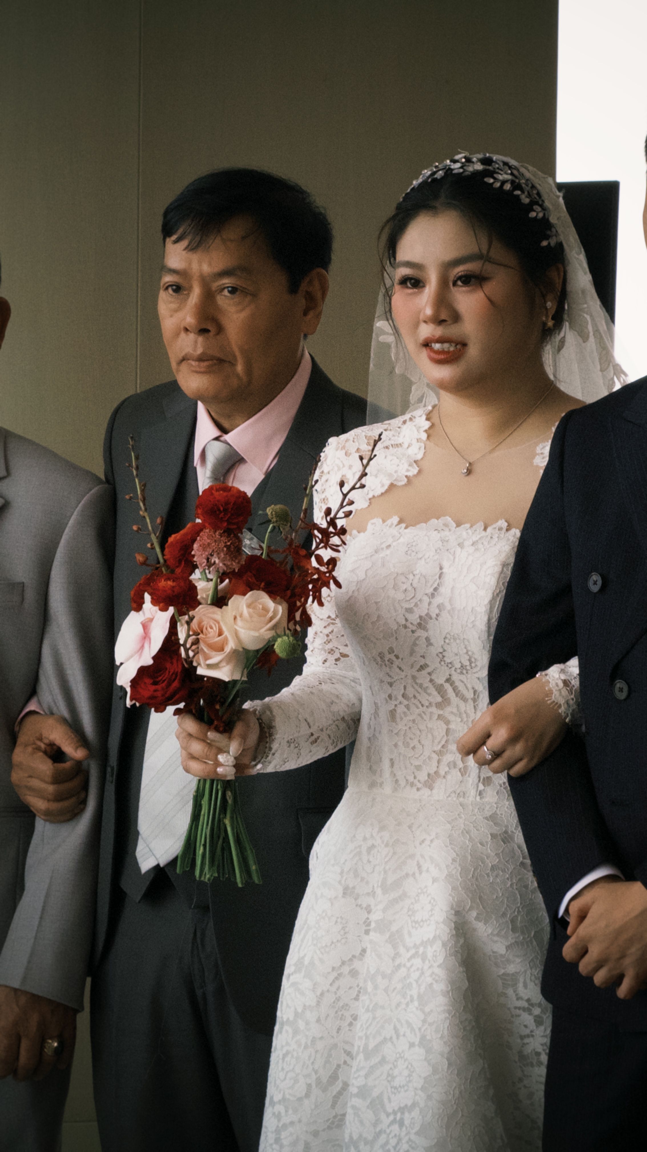 A Vietnamese bride in a white gown holds a bouquet, standing with her family.