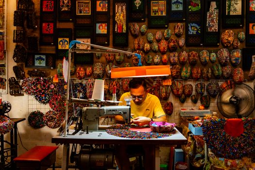 A man works with colorful fabrics in a vibrant handicraft workshop, surrounded by decorative masks and art.