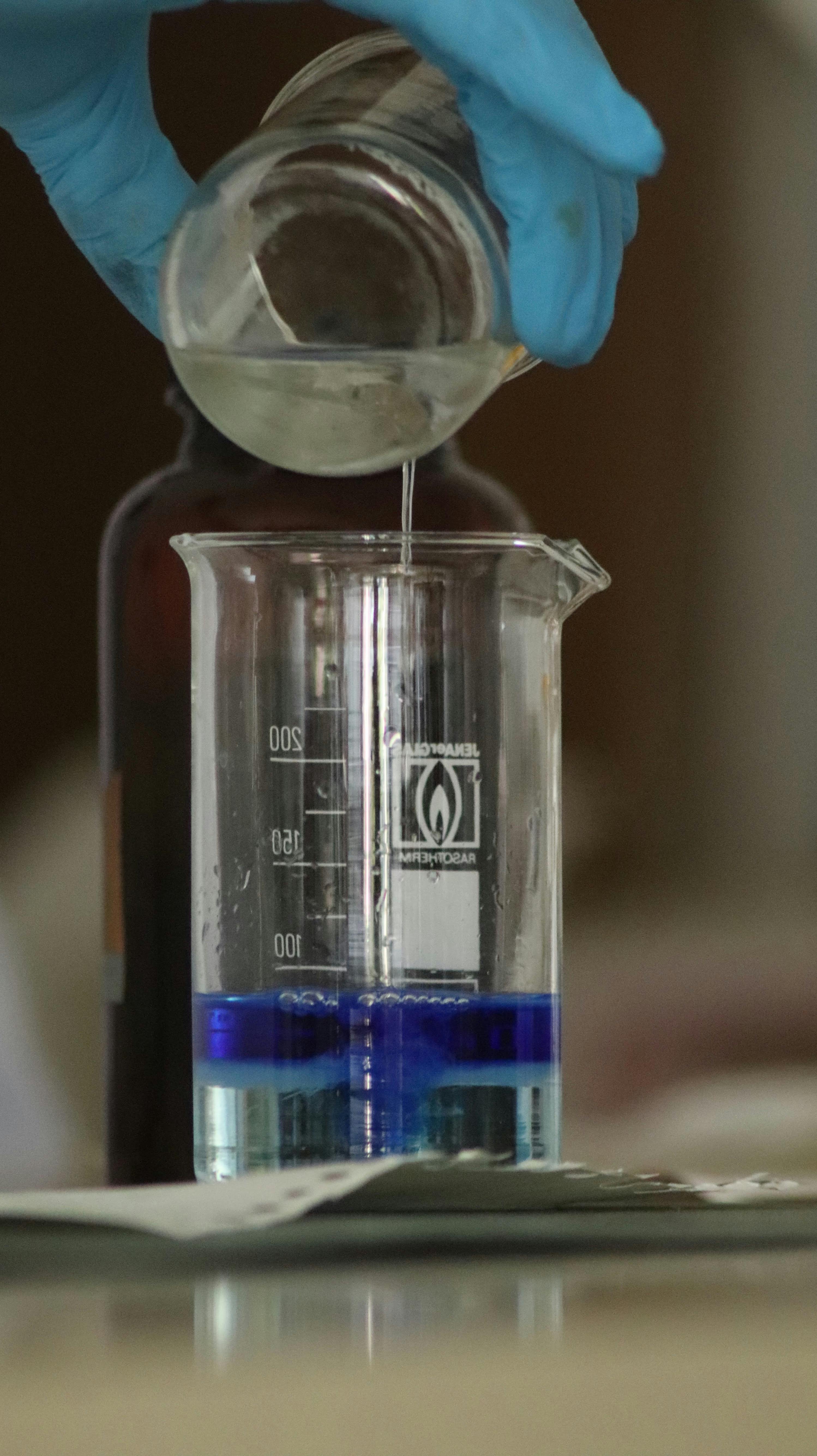 Scientist pouring clear liquid into a beaker with blue solution in a laboratory setting.