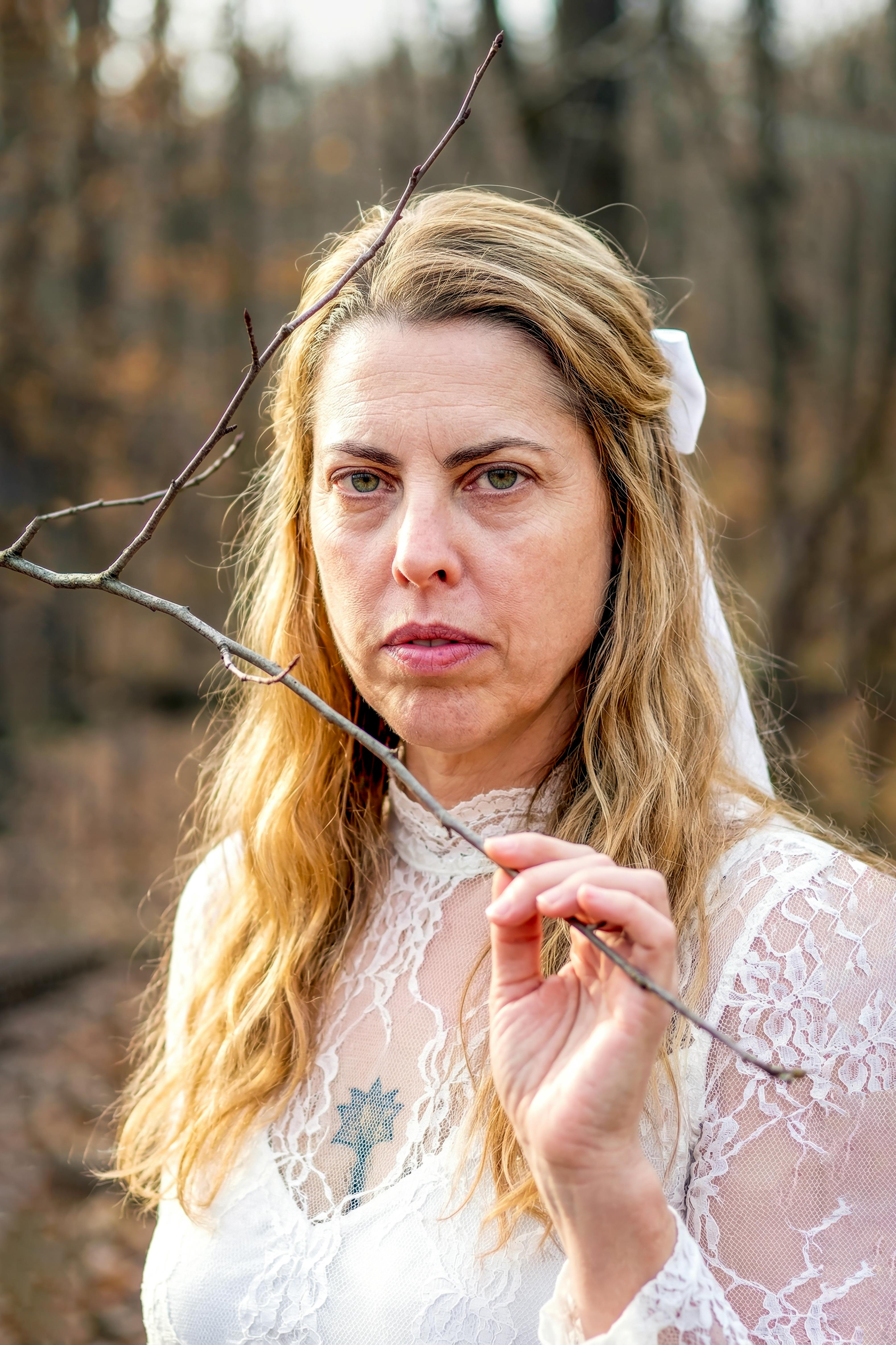 A woman in lace attire holds a twig in a contemplative forest setting.
