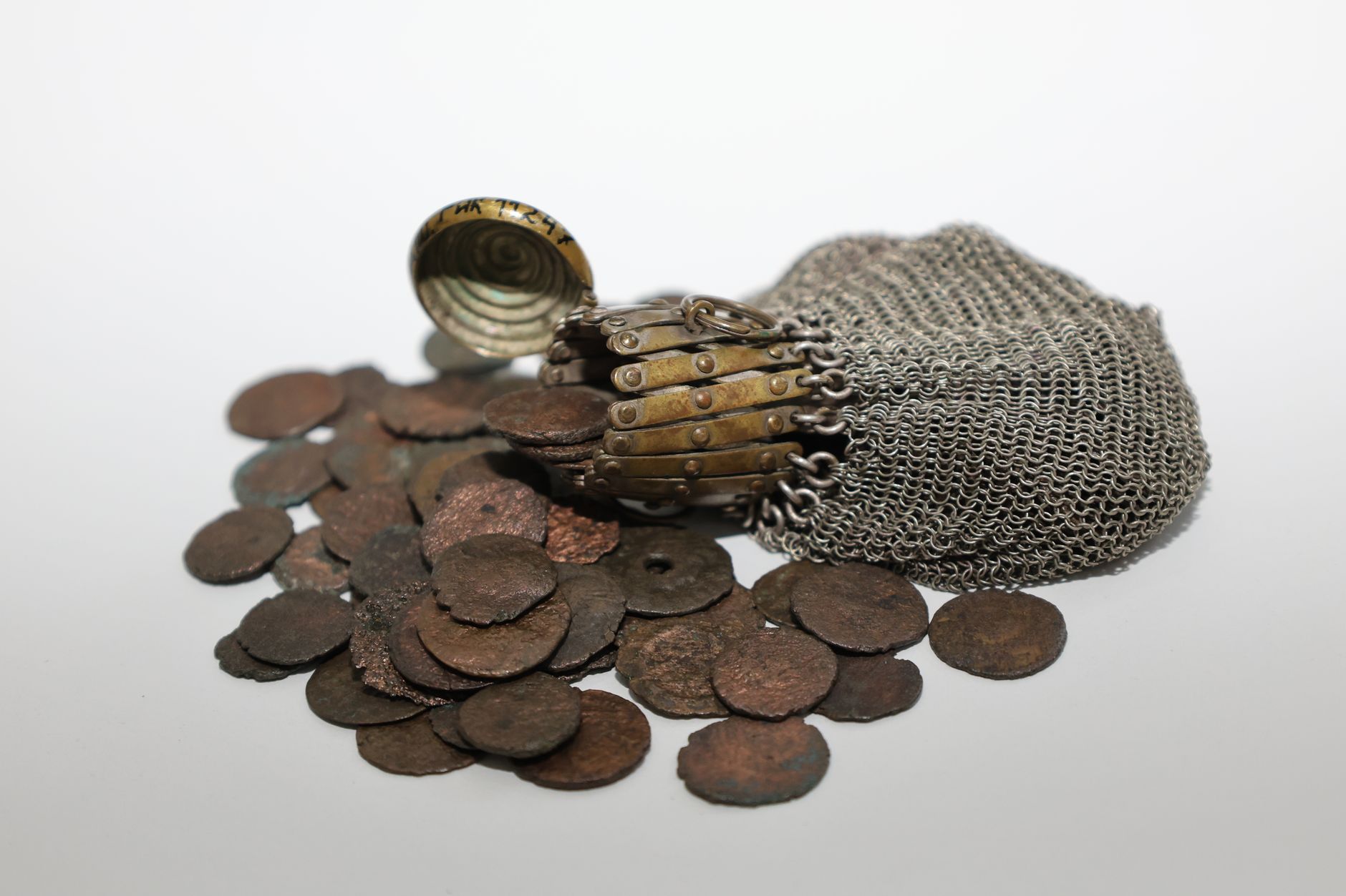 Antique metal chain pouch spilling ancient rustic coins on a white surface.