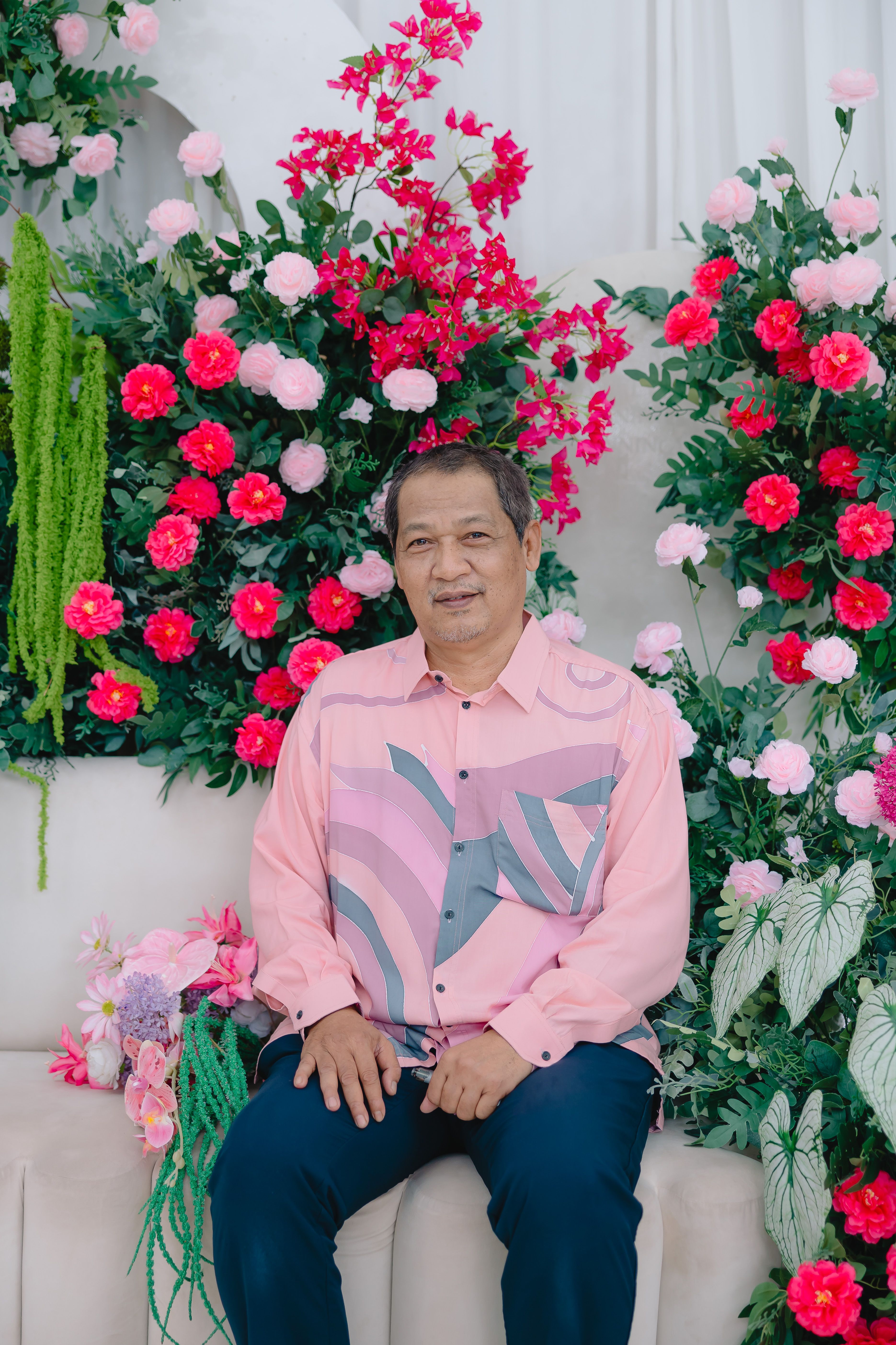 A senior man in a pastel shirt sits amid a colorful floral arrangement, exuding elegance and warmth.