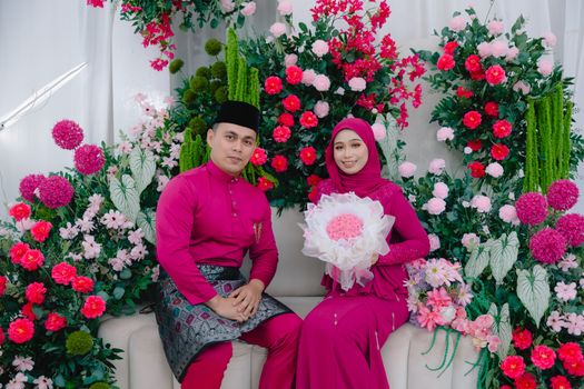 A couple in traditional Malay attire posing amidst colorful floral arrangements at a wedding.