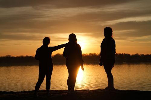 Free Three friends silhouette against a stunning sunset by a tranquil lake. Stock Photo