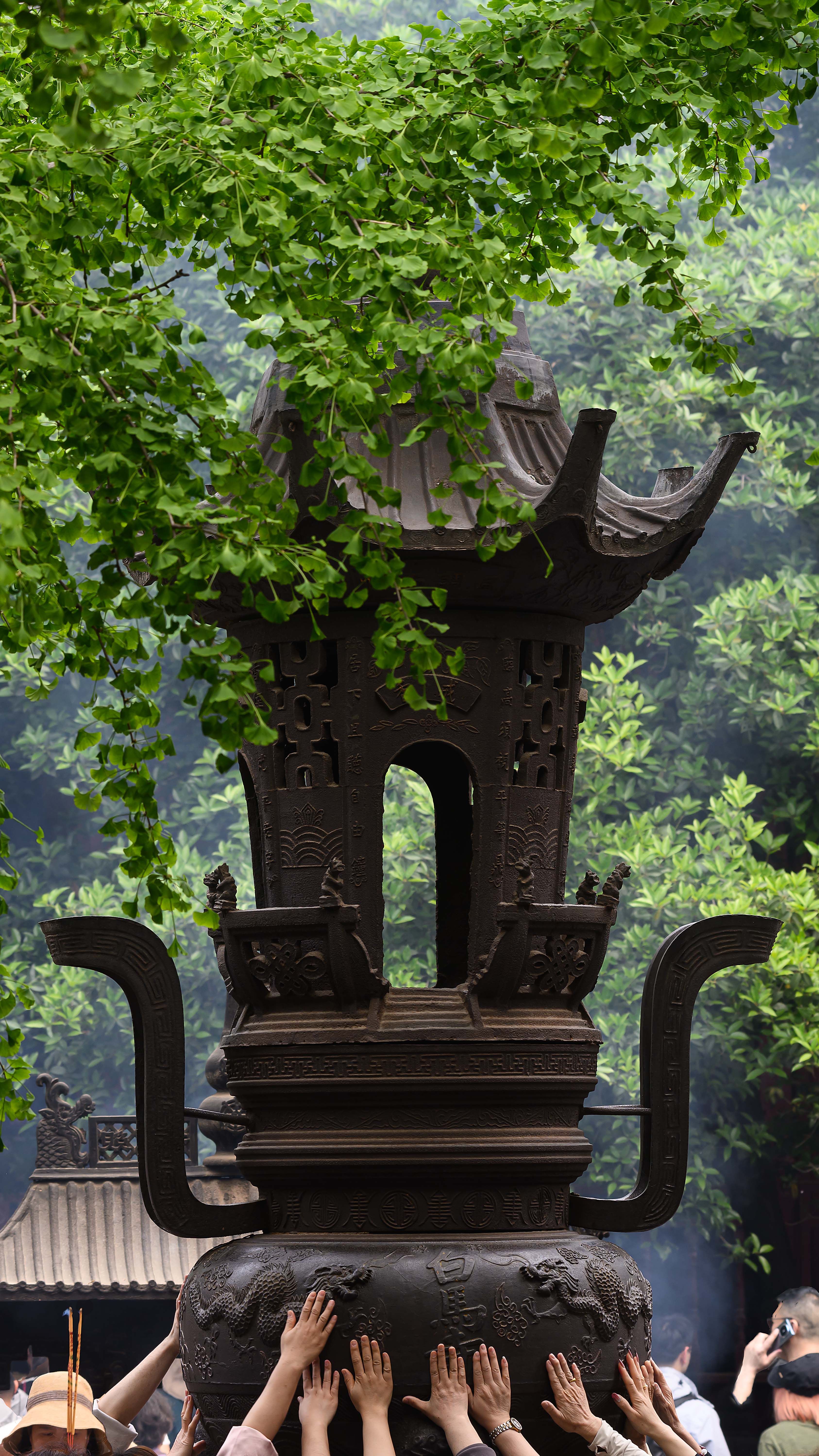 People reaching for a traditional temple incense burner surrounded by lush greenery on a foggy day.
