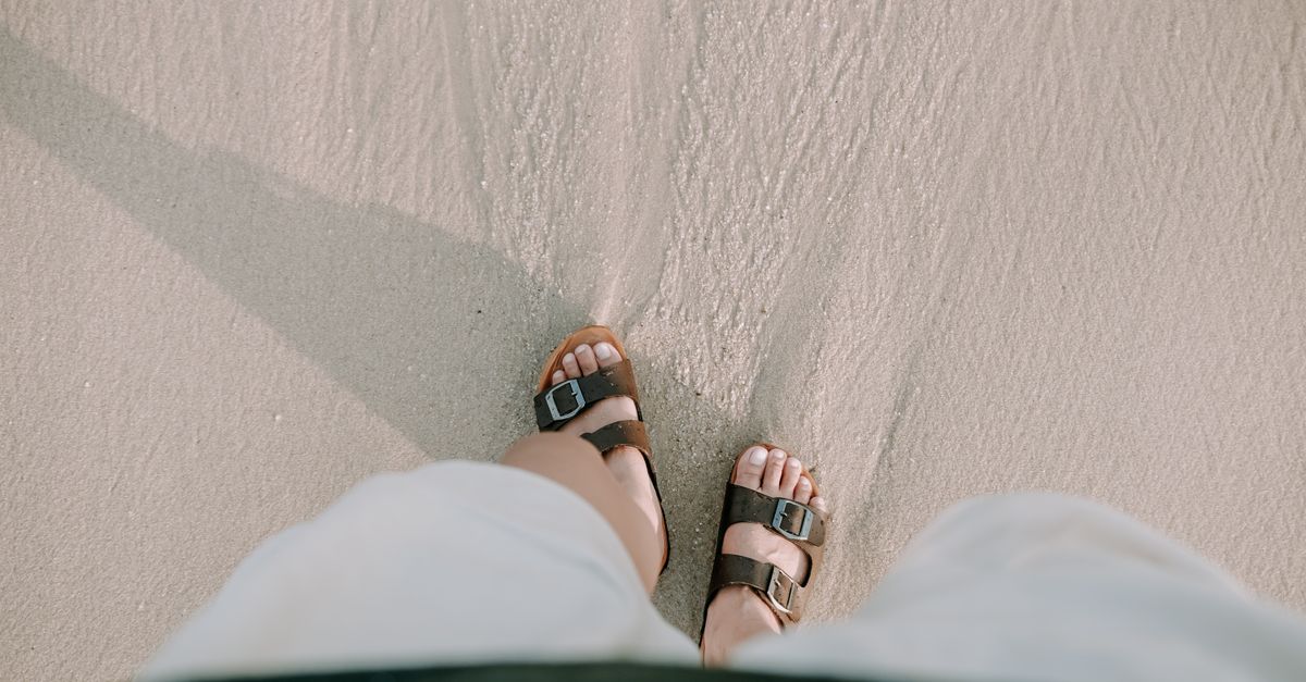 Close-up of feet in sandals strolling on a sandy beach, capturing a serene summer vibe.