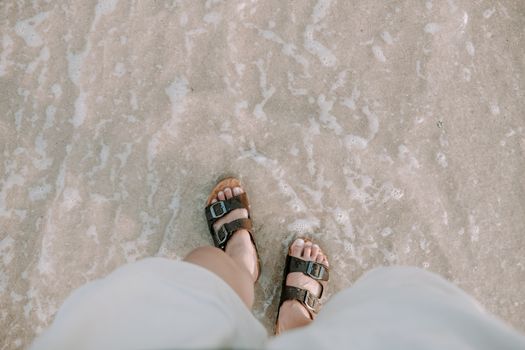 Close-up of feet in sandals standing on sandy beach shoreline with gentle ocean waves.