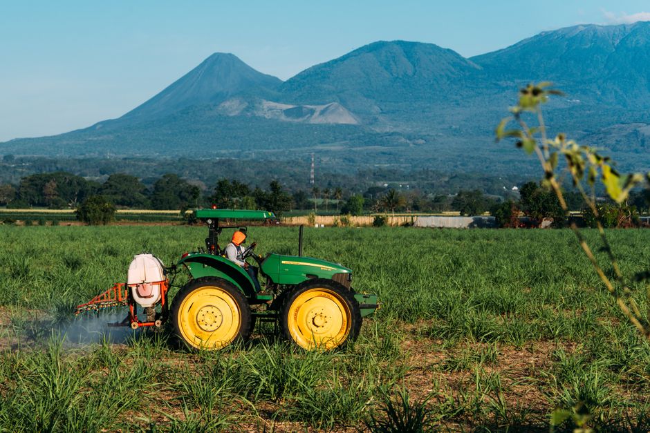 Photo by Edu Raw A farmer operates a green tractor in a lush field with a scenic mountain backdrop.