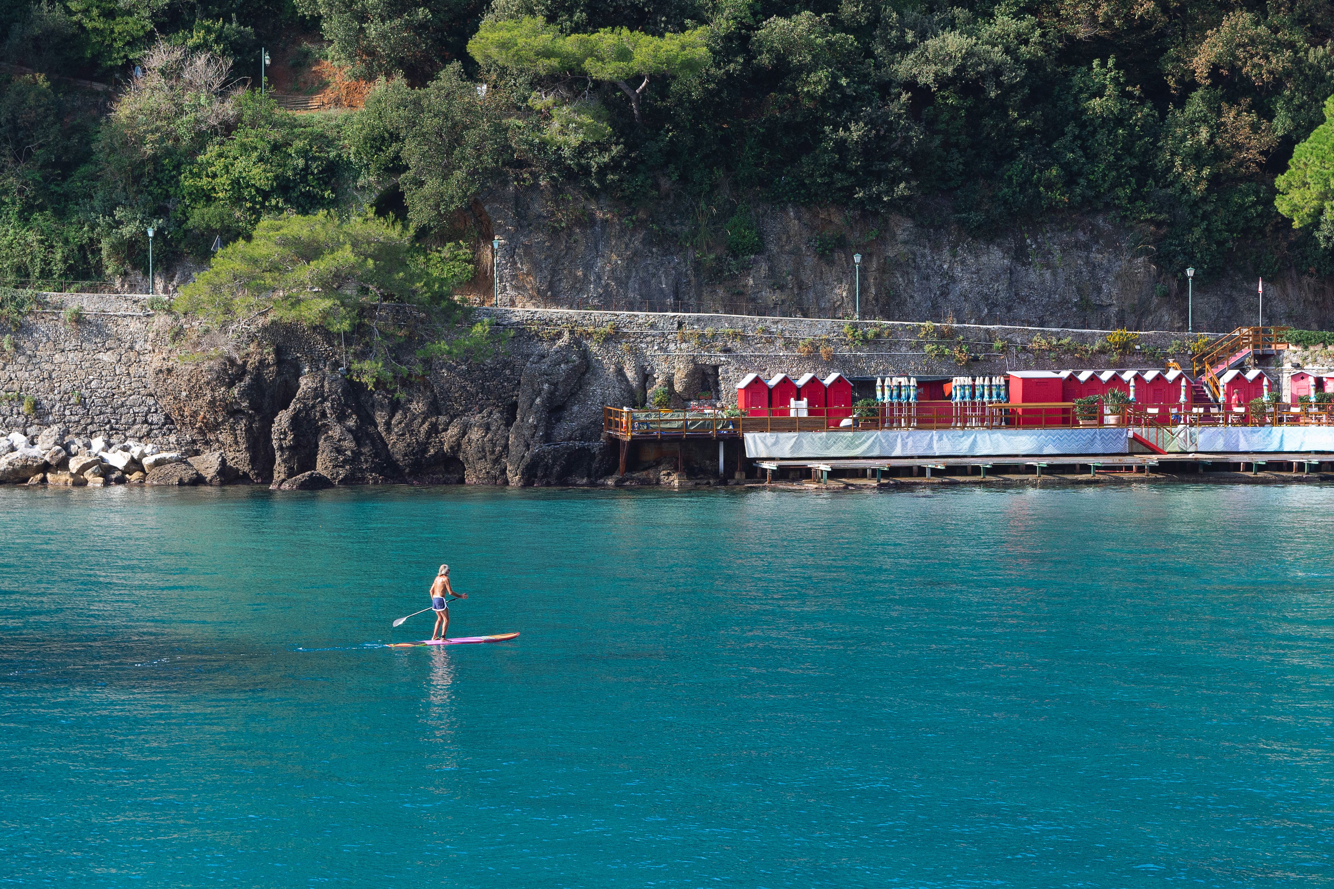 A person paddleboarding on clear turquoise water near a rocky cliff and beach huts.