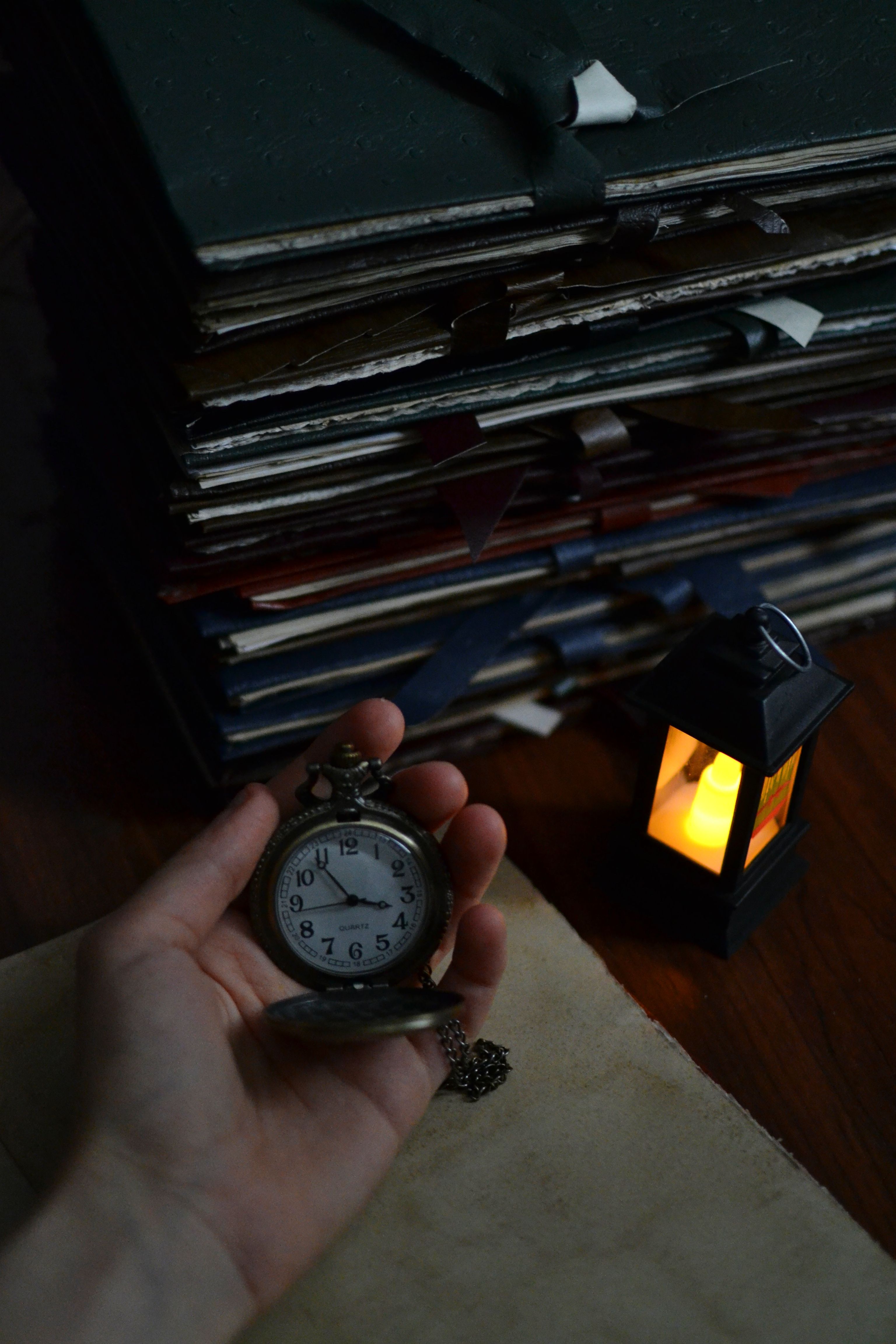 A vintage pocket watch held in hand with a glowing lantern and stack of books in dim light.