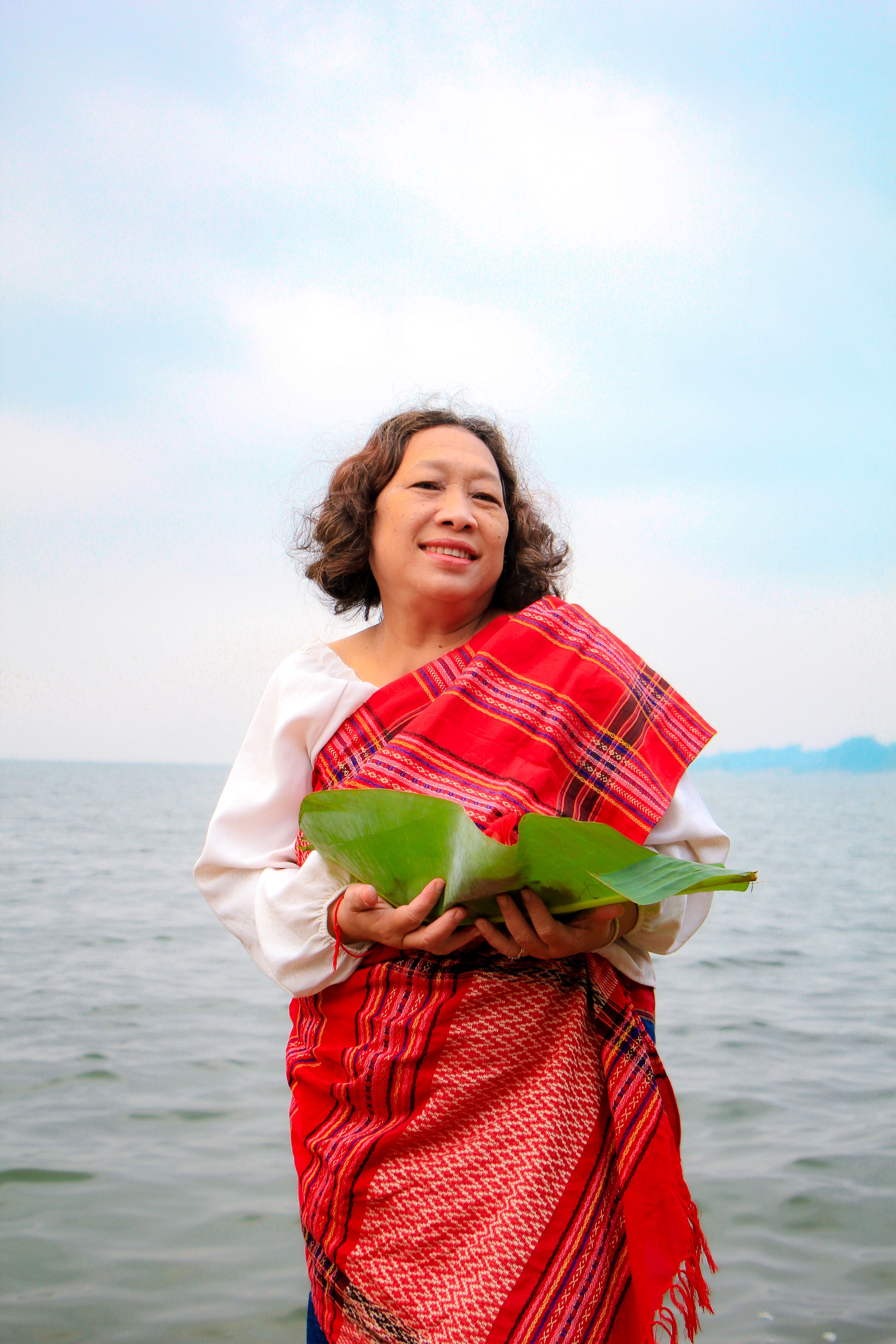 A woman in traditional attire stands by the lake holding green leaves, celebrating cultural heritage.