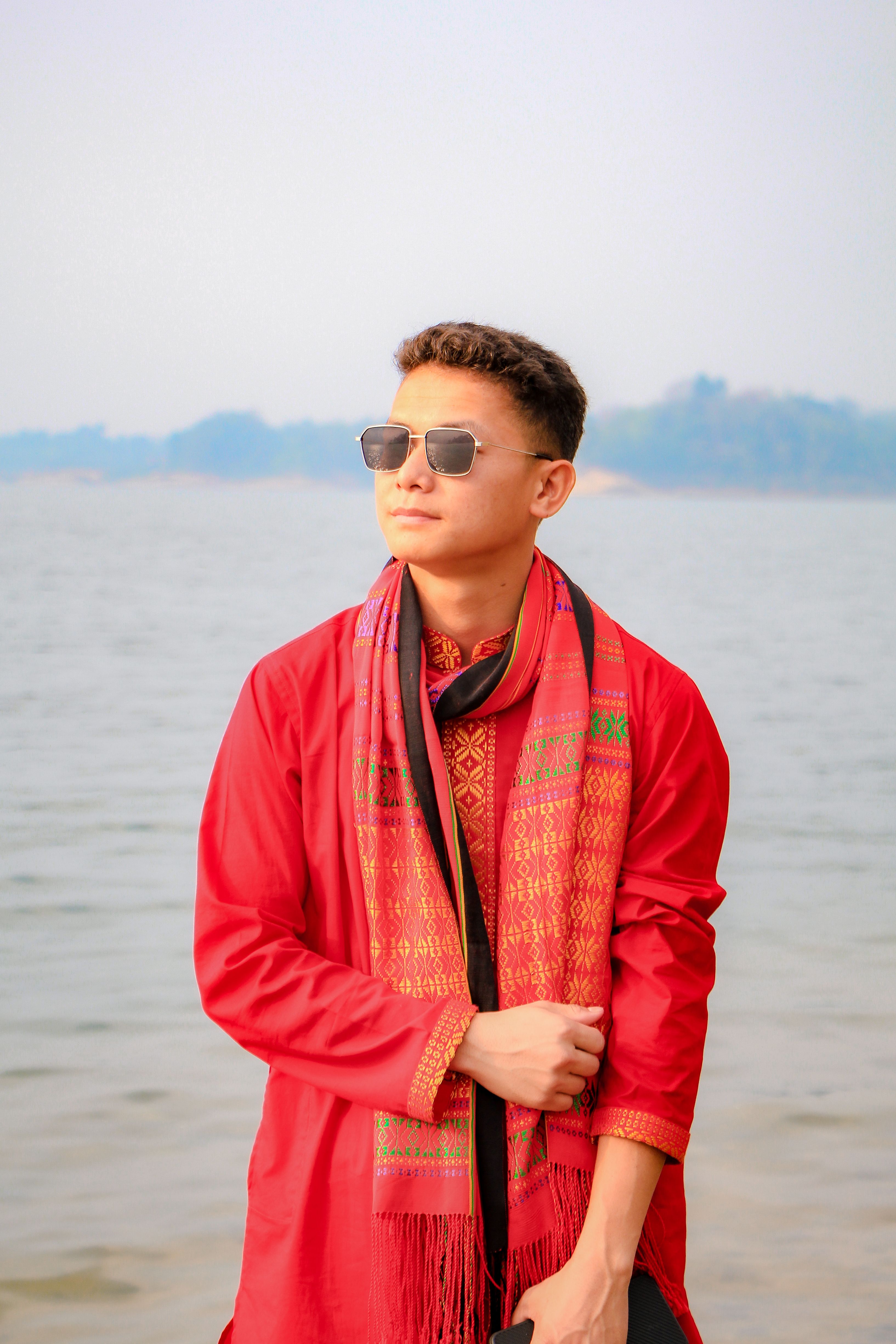 A young man wearing traditional attire stands elegantly by a serene lake.