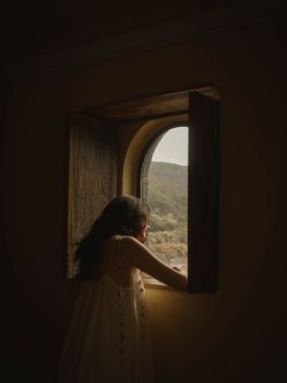 An adult woman looking out a rustic window at nature's landscape, creating a warm, introspective scene.