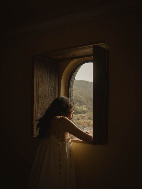 An adult woman looking out a rustic window at nature's landscape, creating a warm, introspective scene.