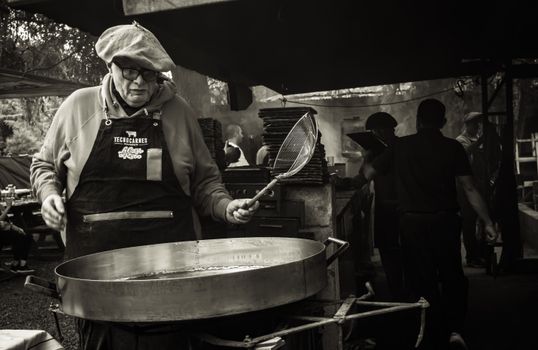 A middle-aged man cooks at an outdoor market stall using a large pan, creating street food in a lively setting.