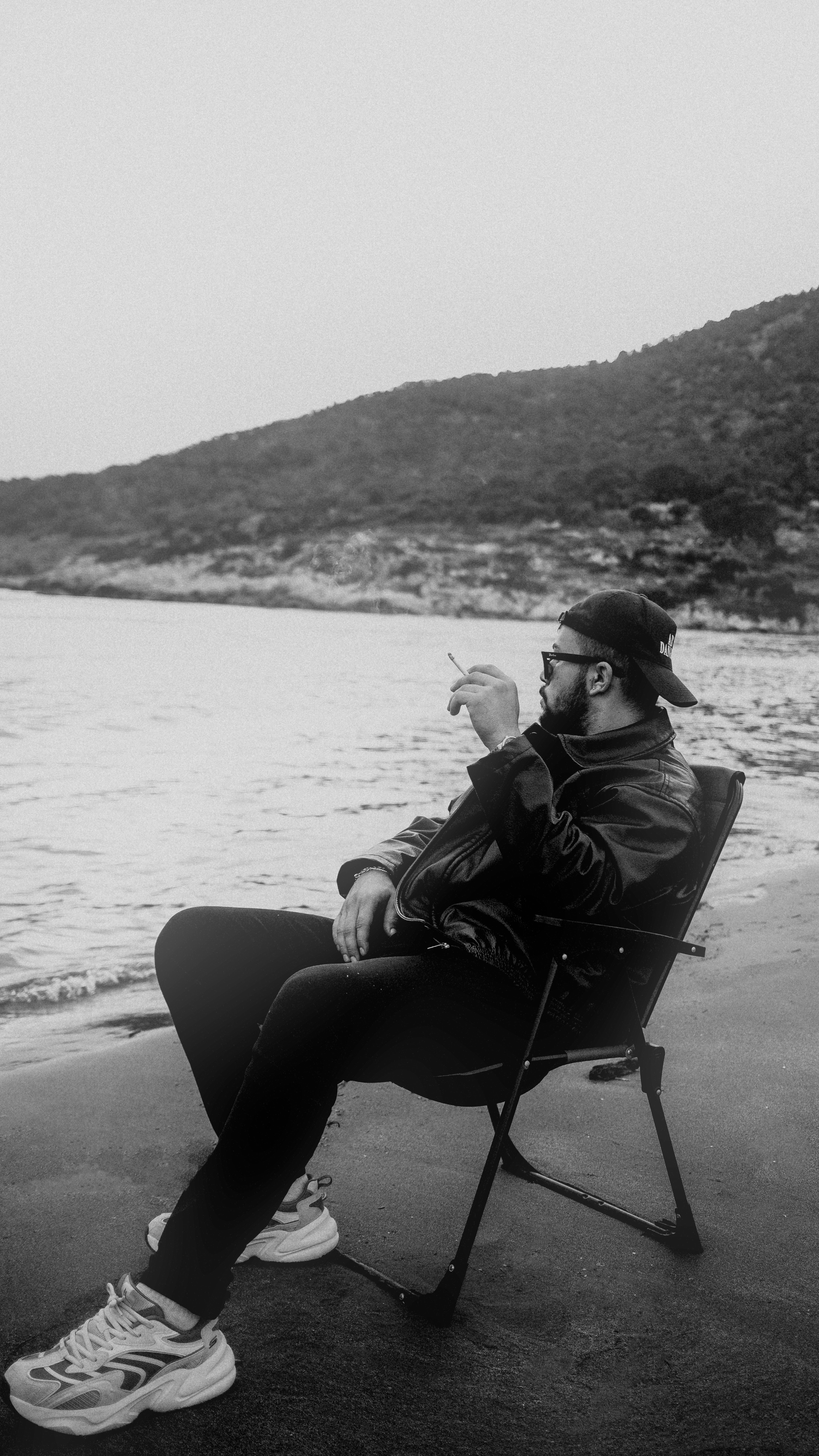 Man in sunglasses and cap smoking in a chair by the ocean, calm setting.
