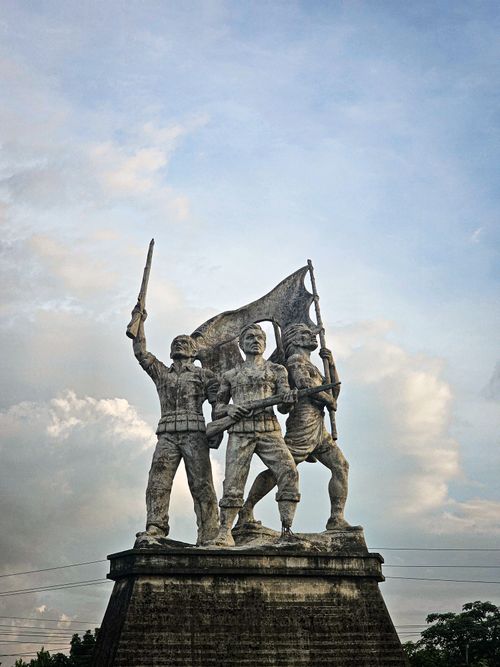 Free Monument of three soldiers holding a flag, symbolizing revolution, with a cloudy sky backdrop. Stock Photo