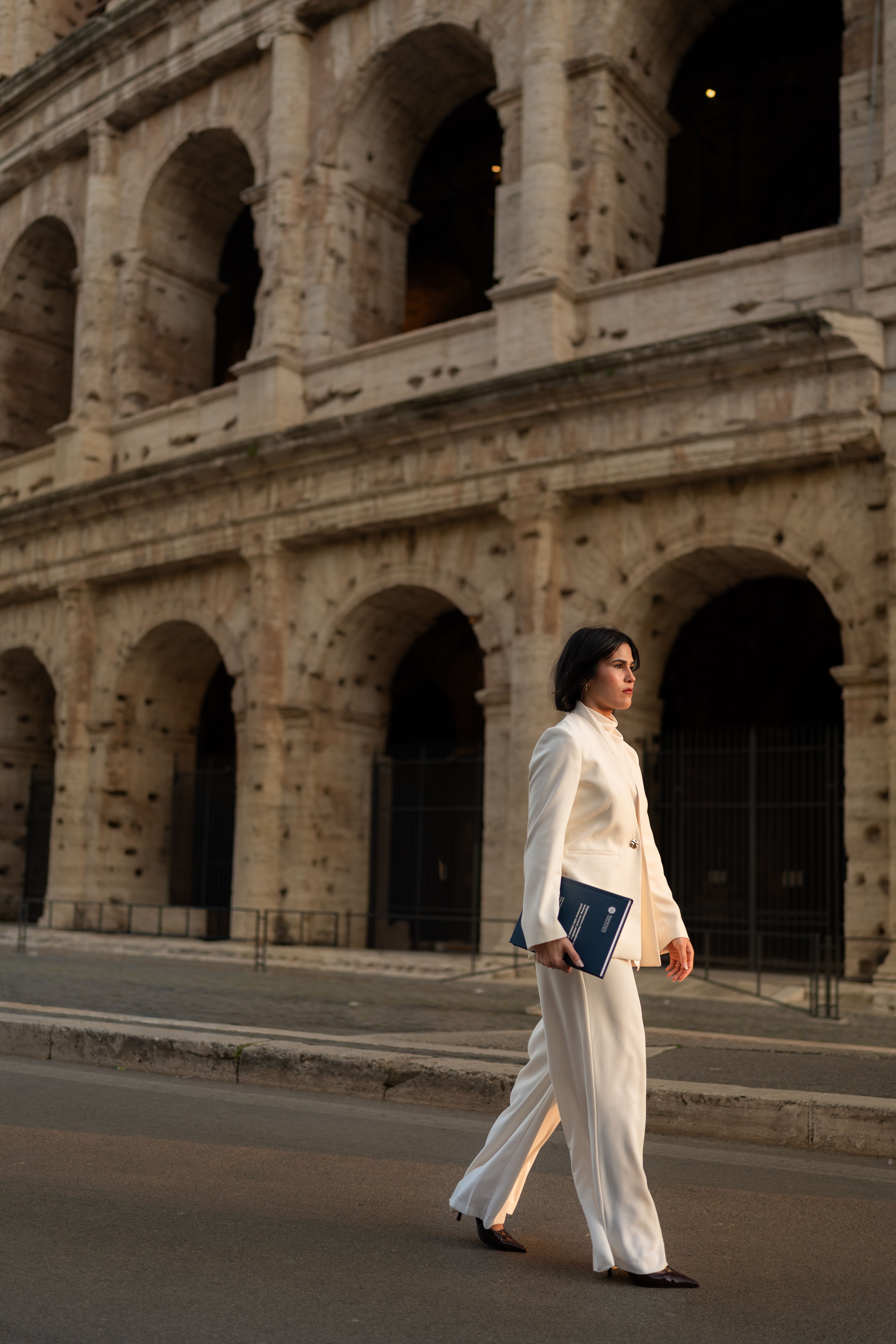 Professional woman in white suit walking past the iconic Colosseum in Rome at dusk.