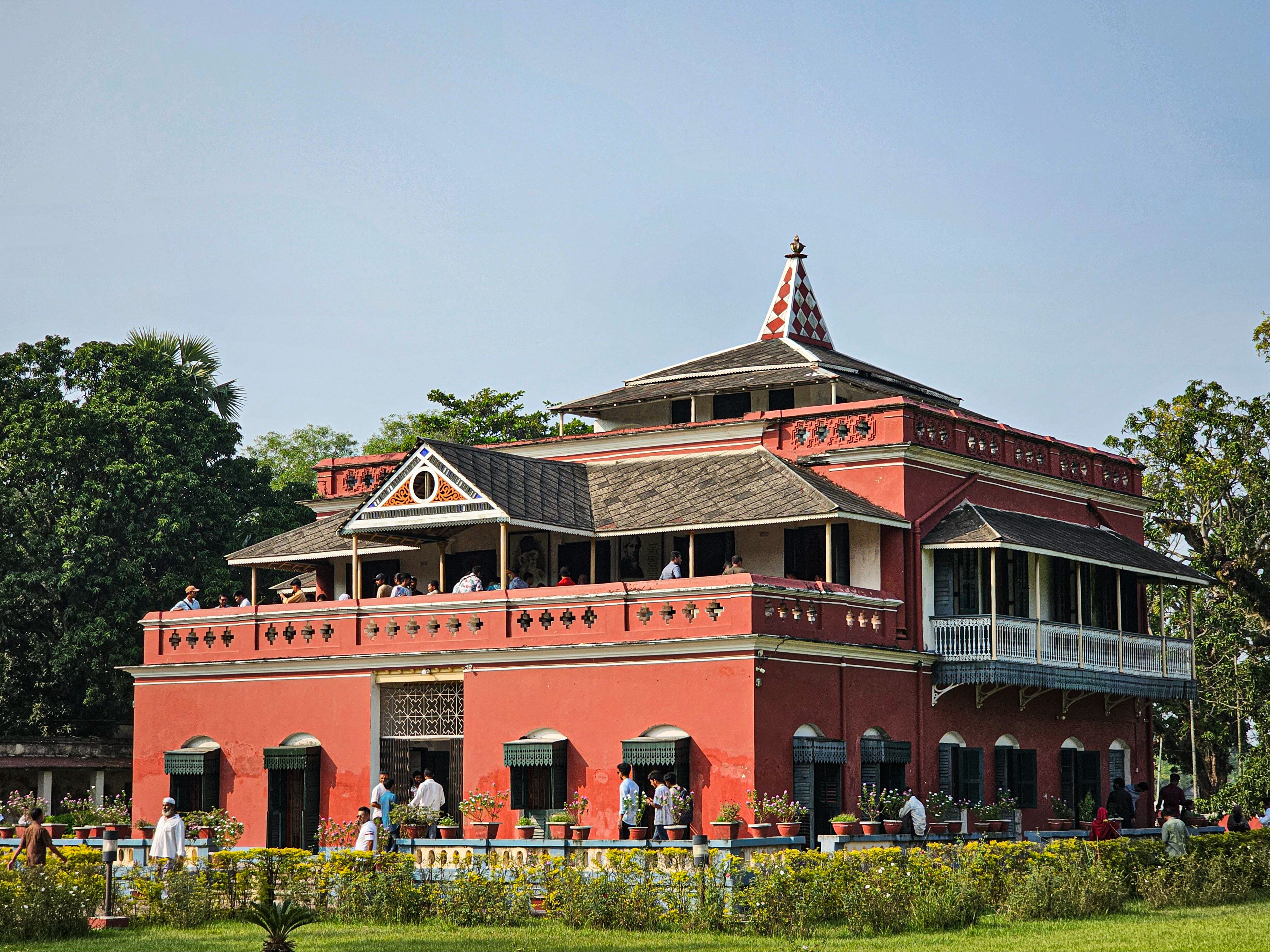 Tourists exploring a historic red building amidst lush greenery on a sunny day.