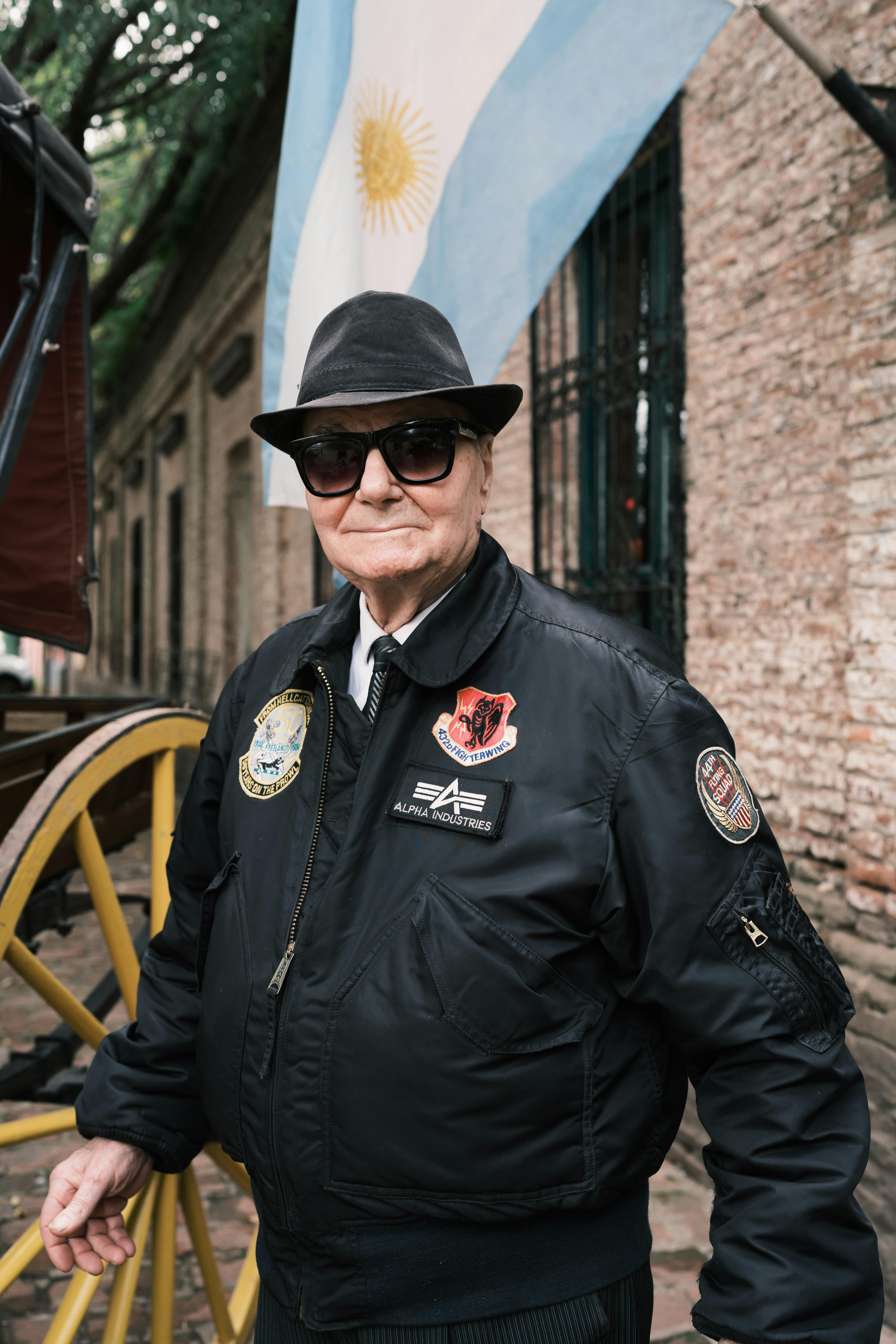 Elderly man in stylish outfit standing in Uribelarrea, Argentina, with Argentine flag in background.