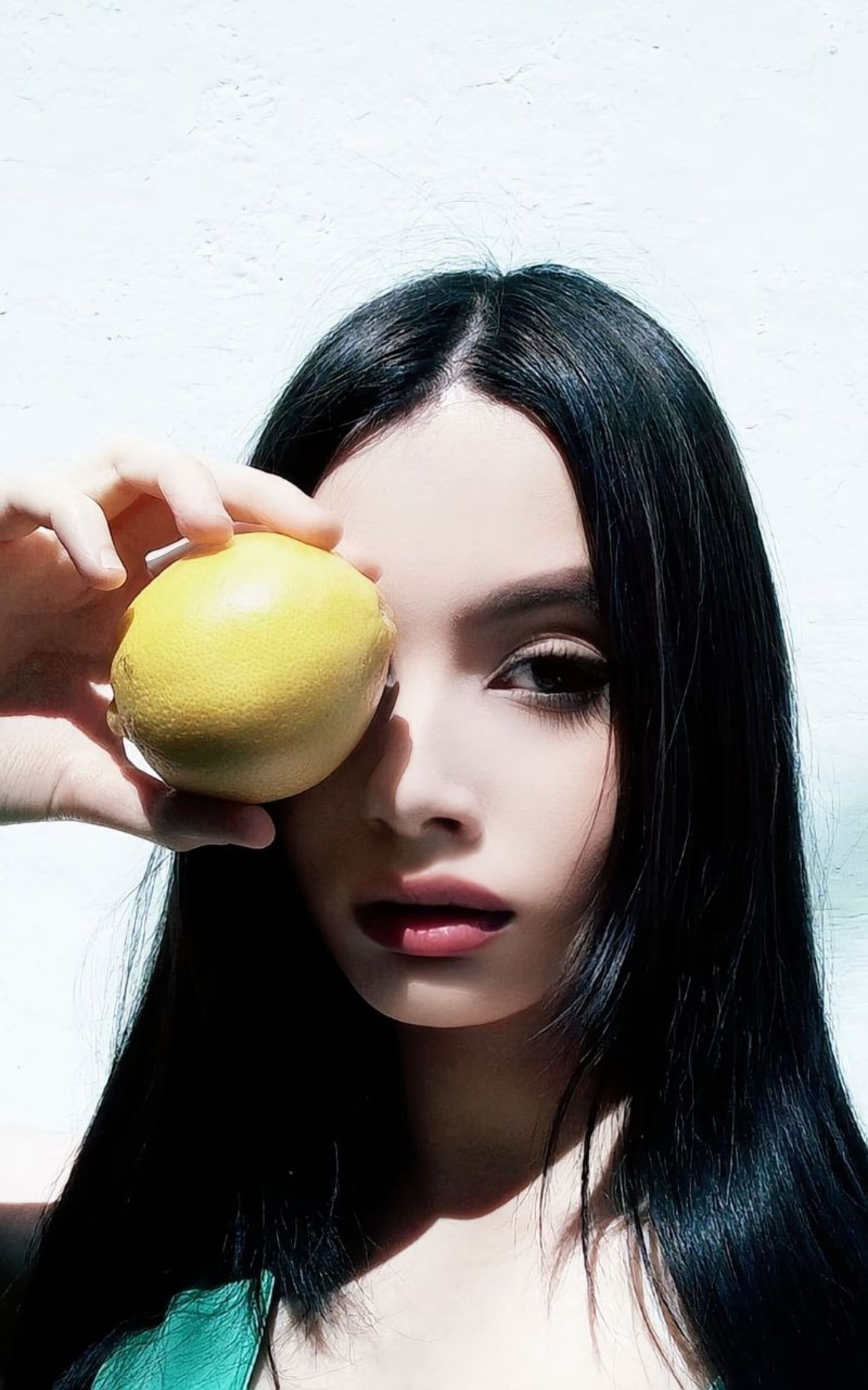 Young woman with long dark hair holding a lemon over her eye in direct sunlight.