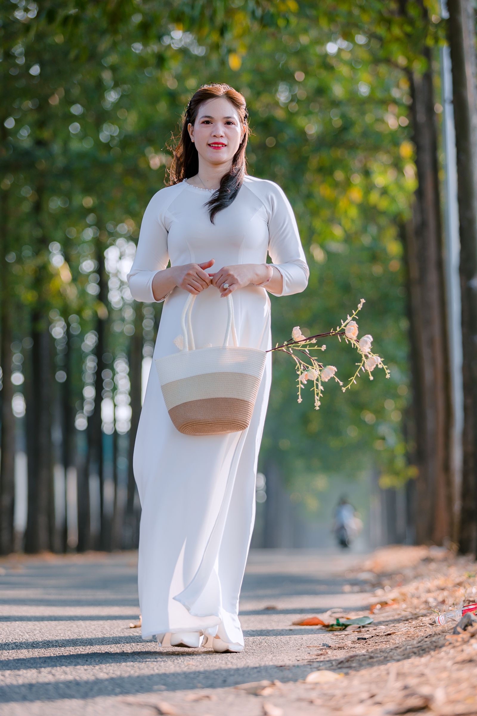 Model in a long white dress holding a woven bag and flowers on a tree-lined path.