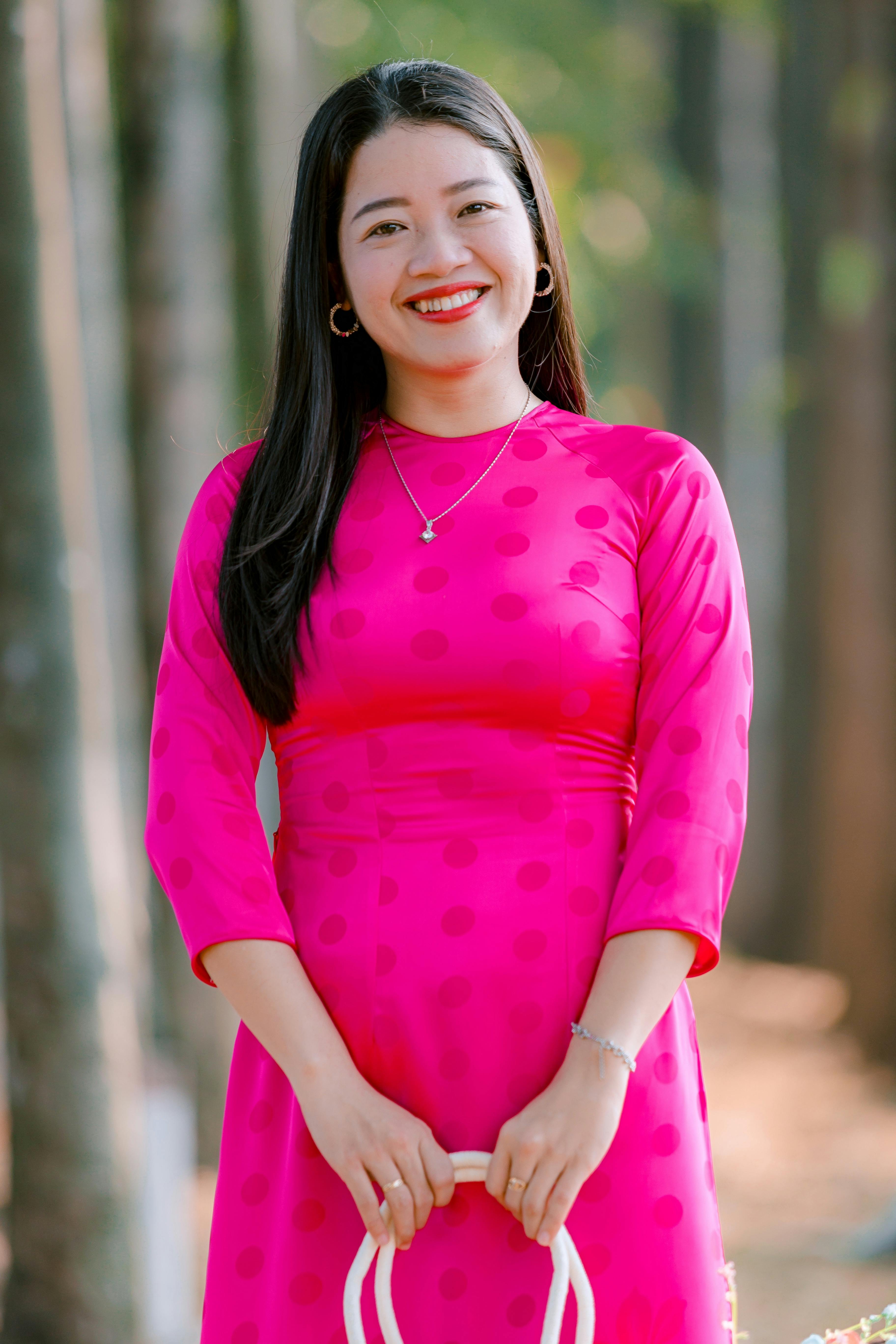 A woman in a vibrant pink dress smiles in a serene forest setting on a sunny day.