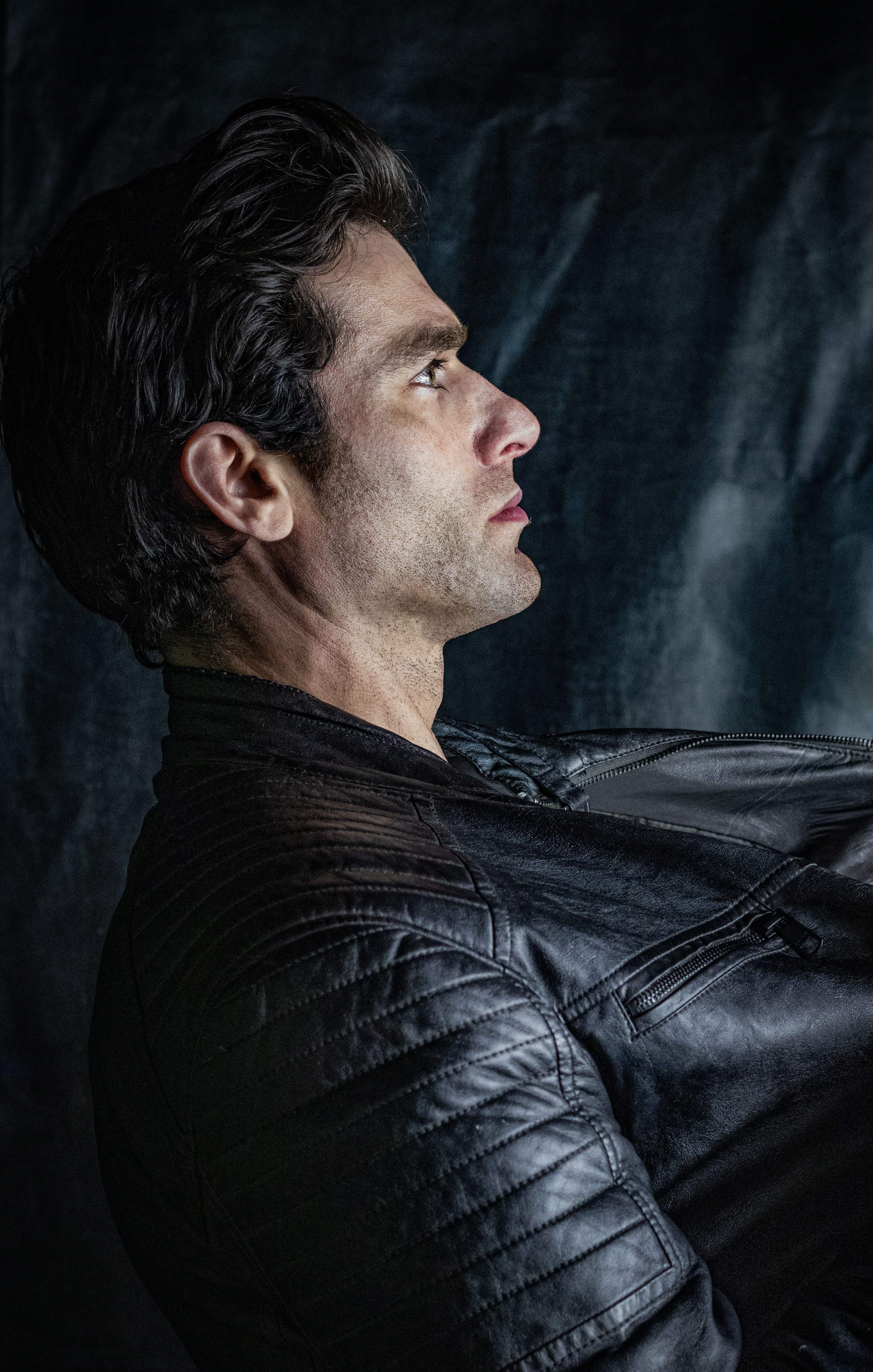 Dramatic side profile portrait of a man wearing a leather jacket against a dark backdrop.