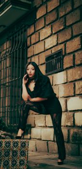 Elegant woman in dark attire poses confidently by a brick wall in urban setting.