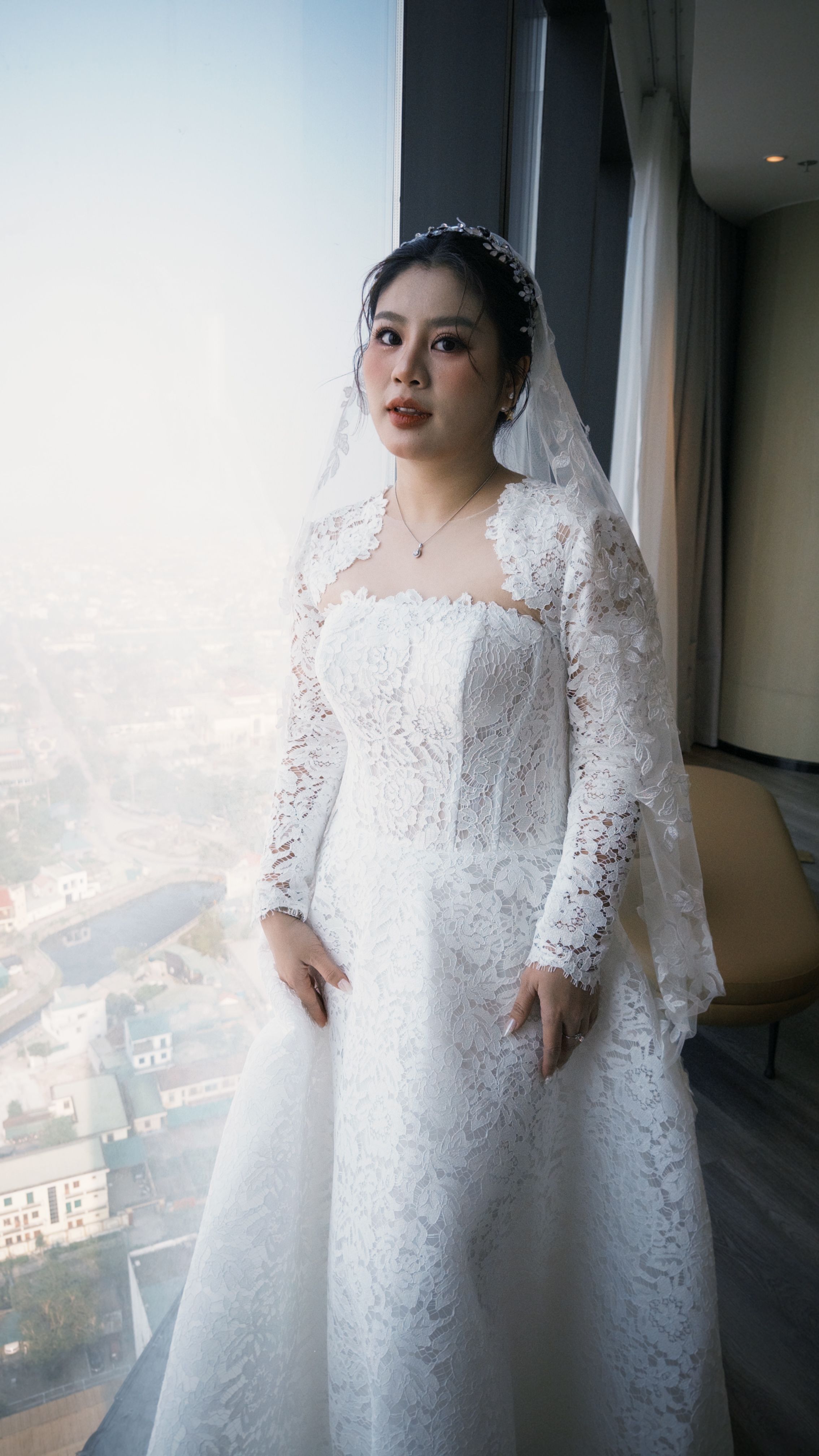 A Vietnamese bride in an elegant lace ao dai, standing by a window with city view.