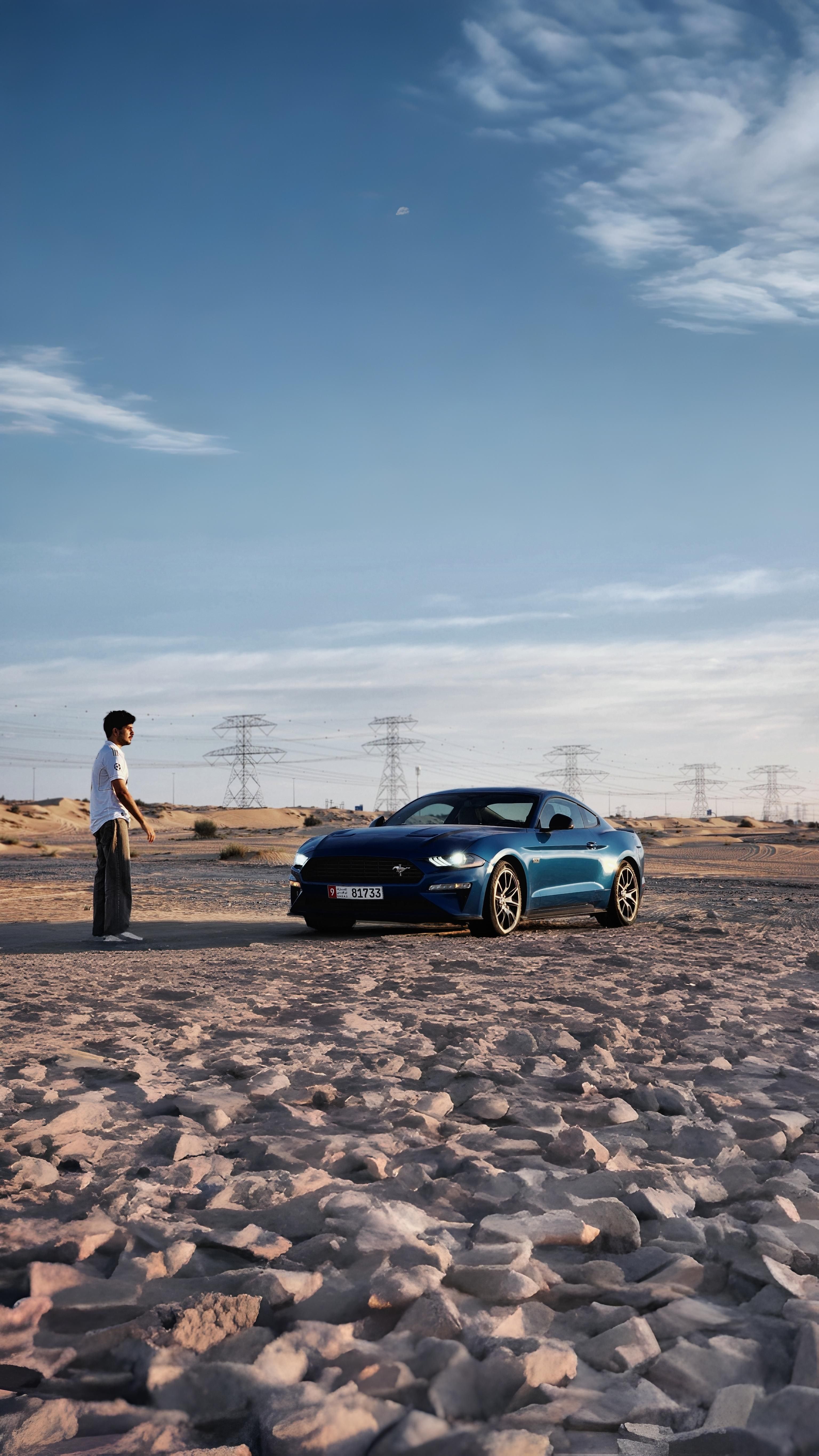 A man stands near a blue sports car in a desert with power lines in the background.