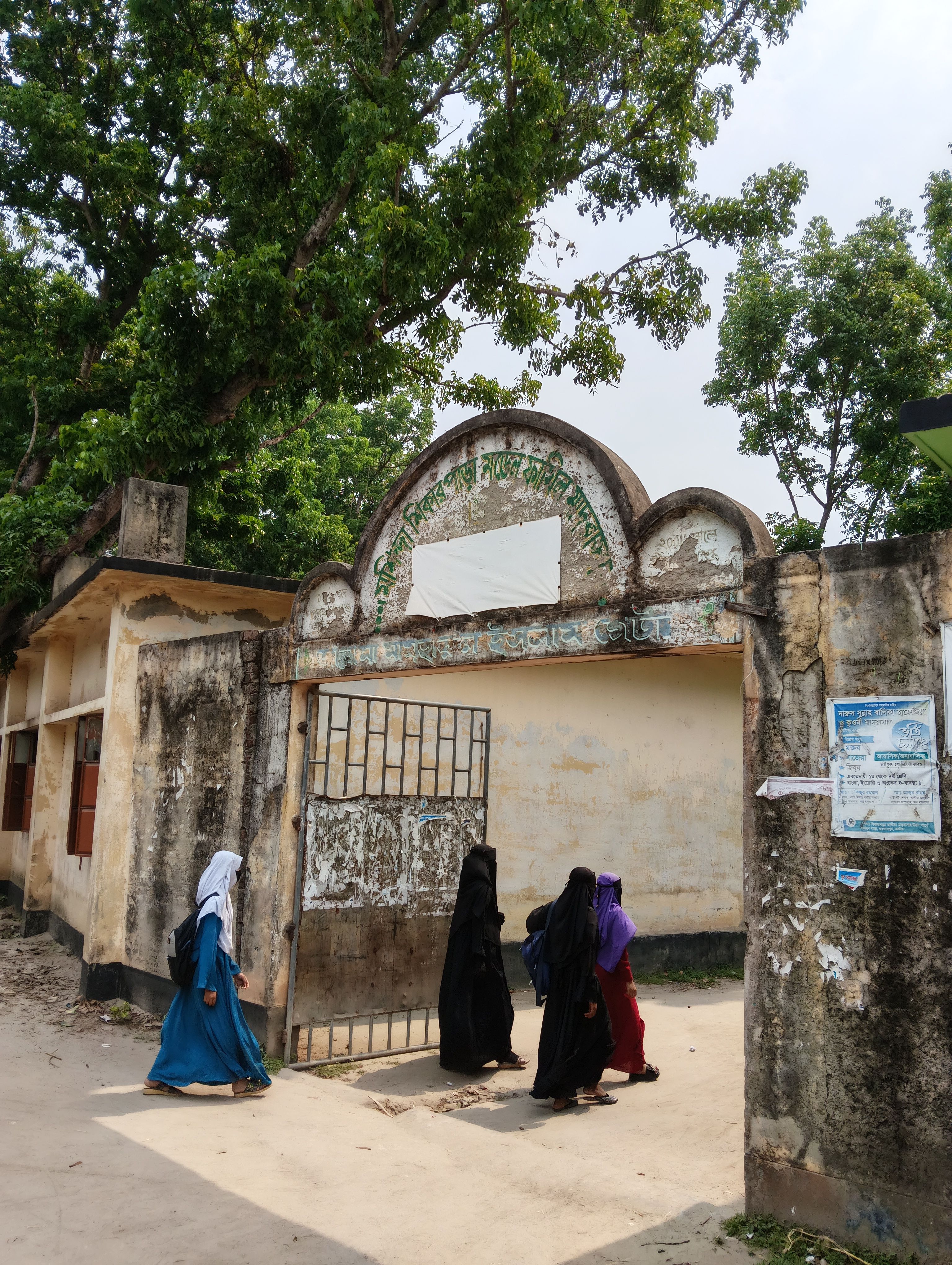 A group of women in colorful traditional outfits walk past a vintage building entrance under lush green trees.