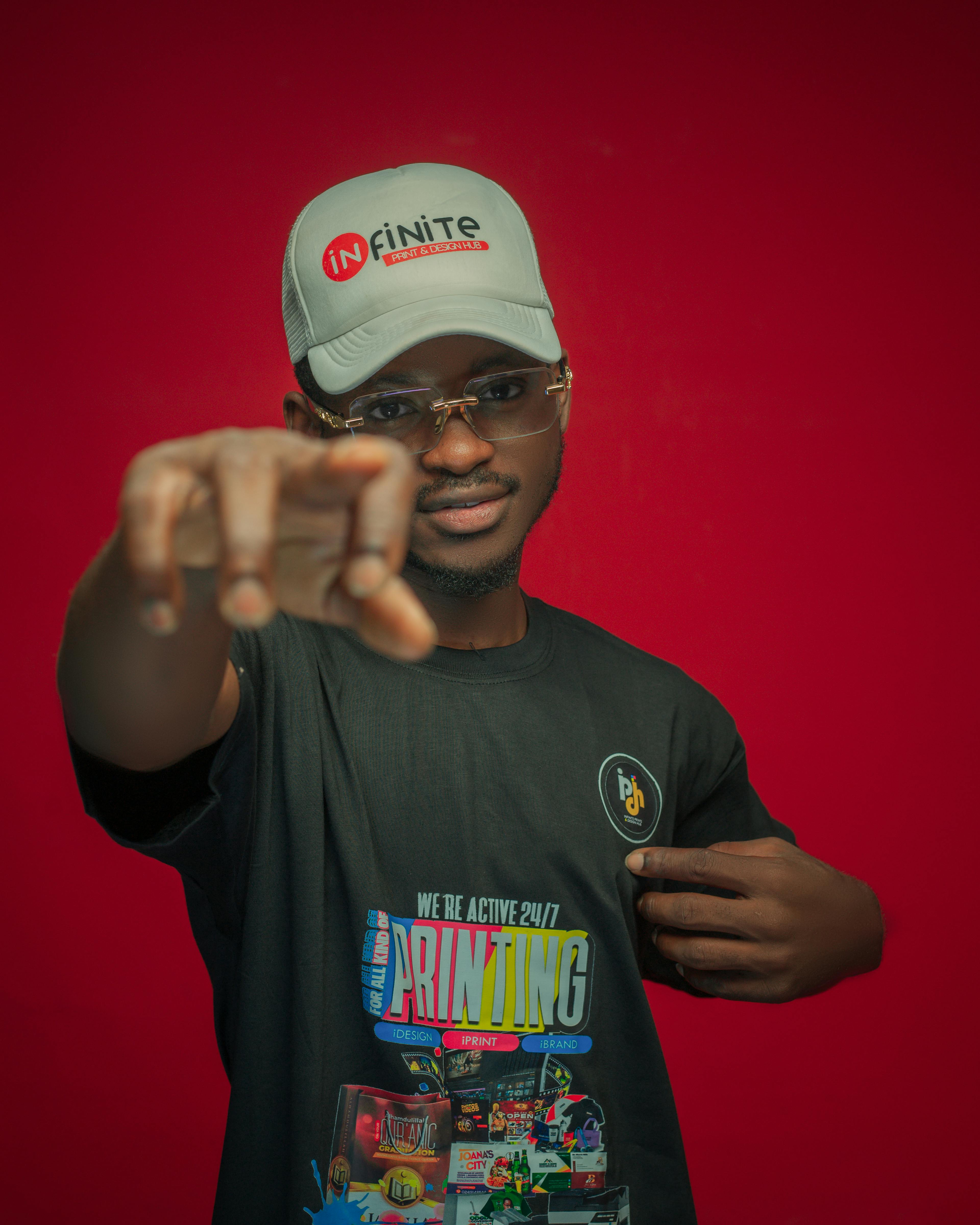 Confident young man wearing a branded cap and t-shirt, pointing directly at the camera against a red background.