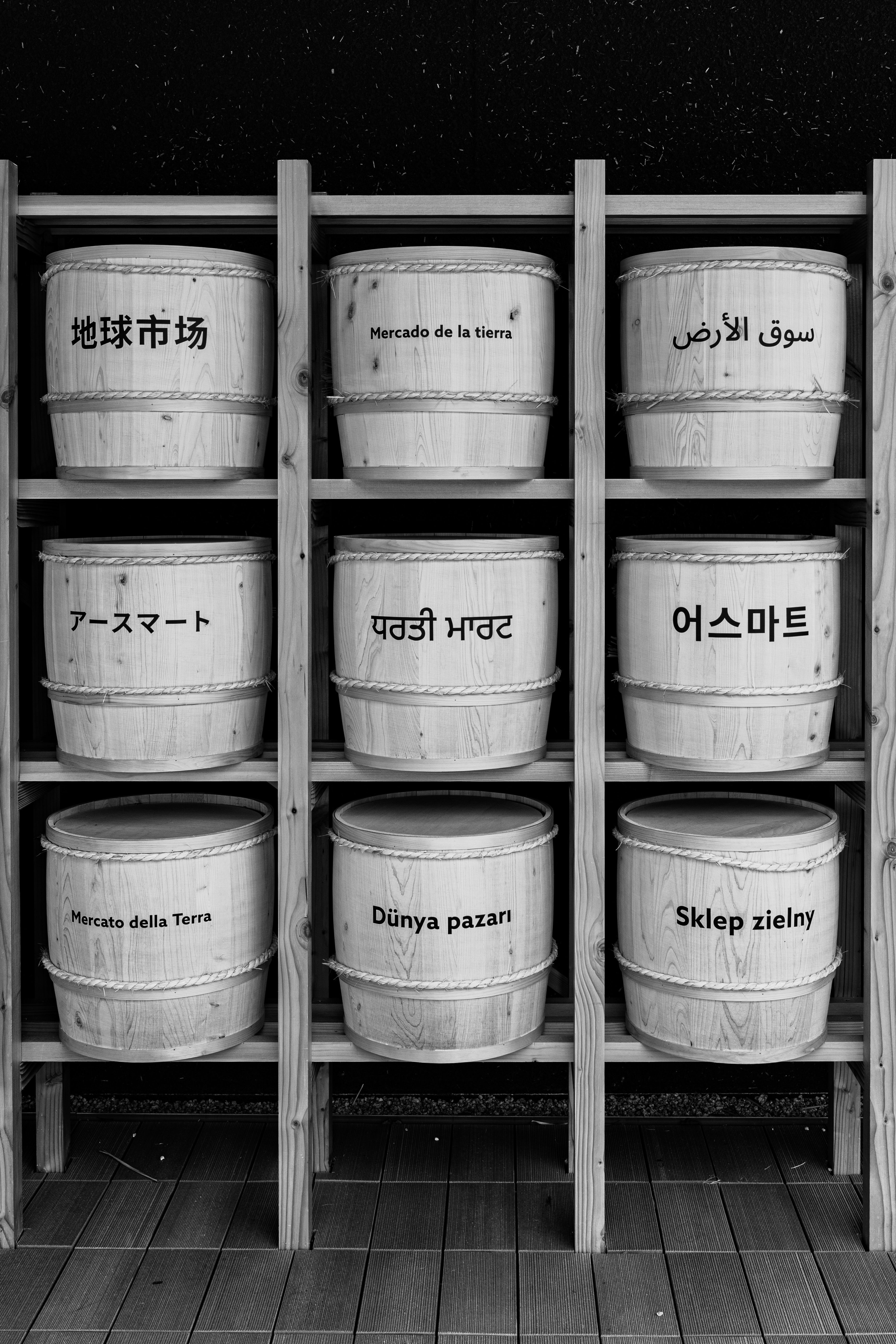 Black and white image of wooden bins with multilingual market names on display shelves.